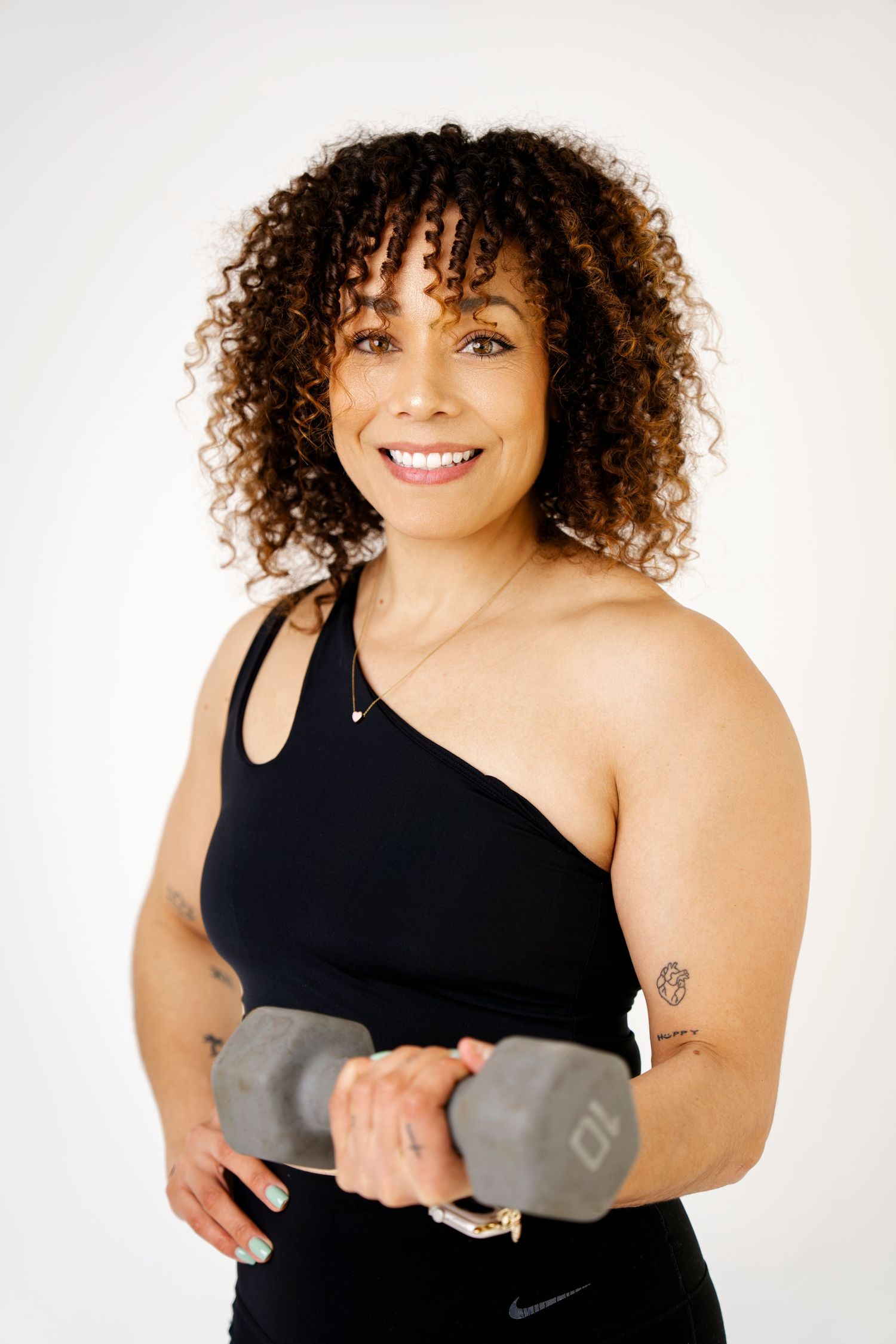 Smiling woman with curly hair holding a dumbbell in black athletic wear against a white background.