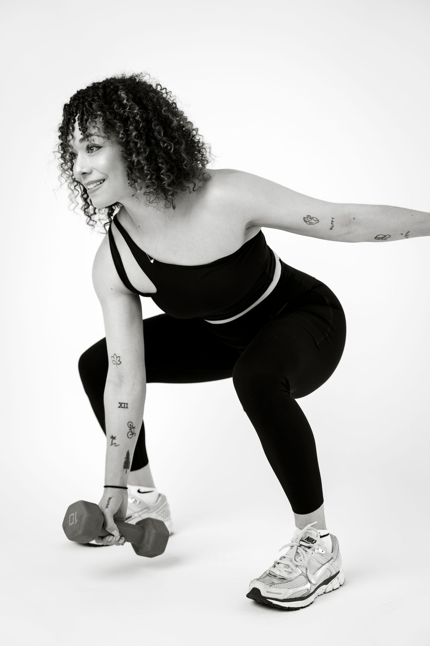 Woman in black athletic wear doing a squat with dumbbells, showcasing fitness form and tattoos on arms.