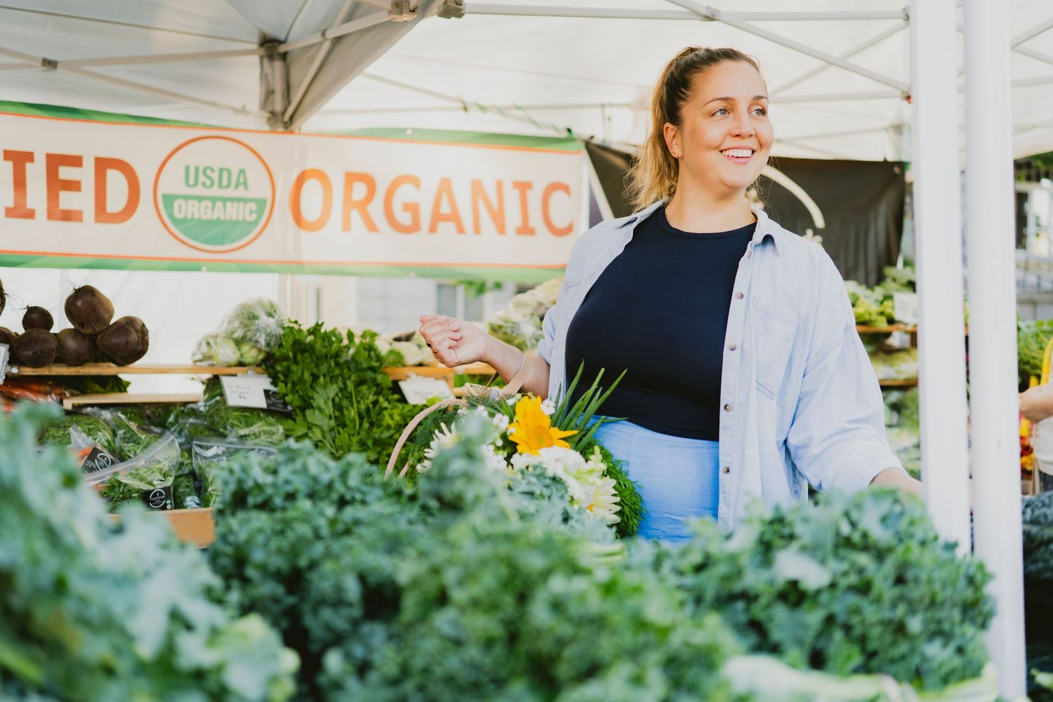 Woman shopping at a USDA certified organic farmers market stand with fresh kale and vegetables.