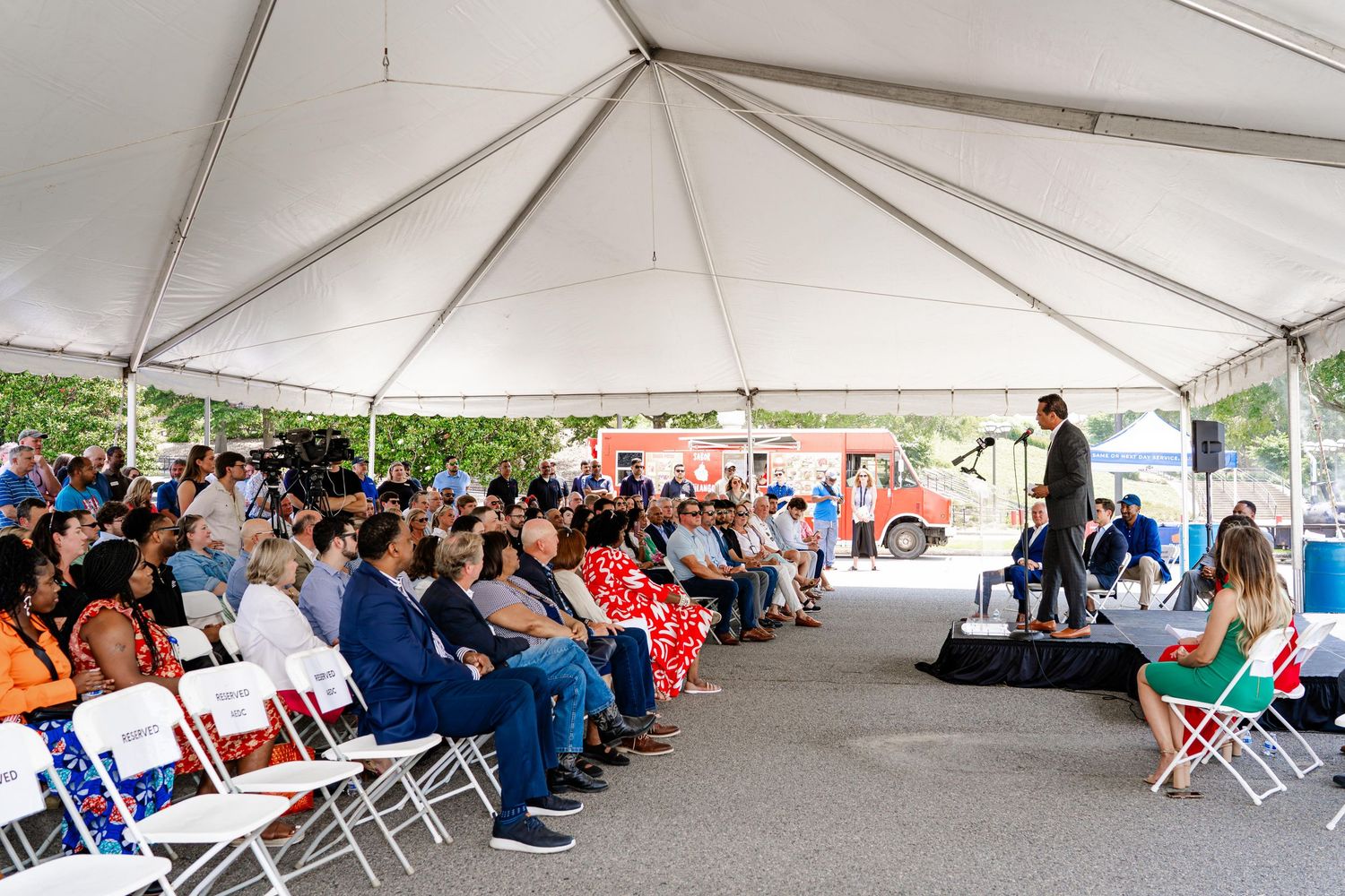Large crowd seated under white tent during outdoor community event.