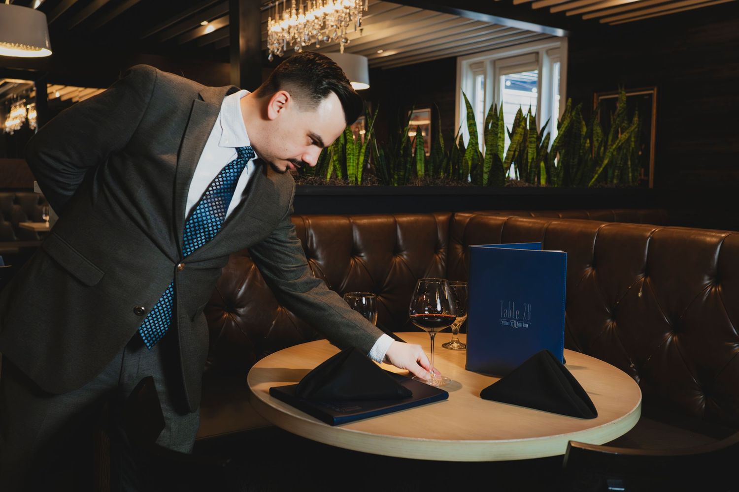 A suited waiter carefully arranges a black napkin on a round wooden table in an elegant upscale restaurant.