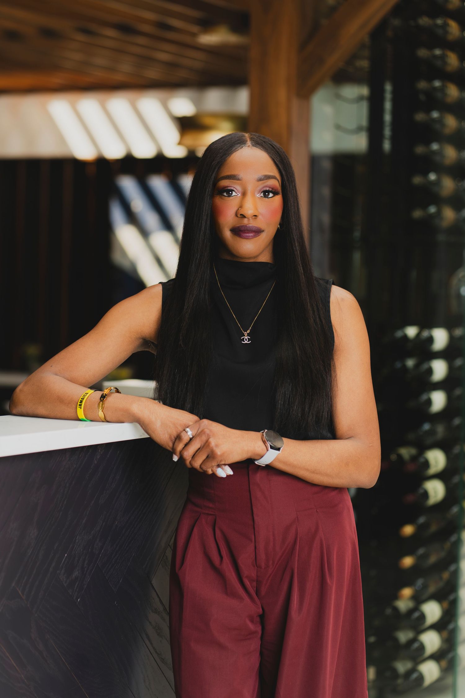 A stylish woman in a black sleeveless turtleneck and burgundy trousers leans against a bar counter in an upscale restaurant.