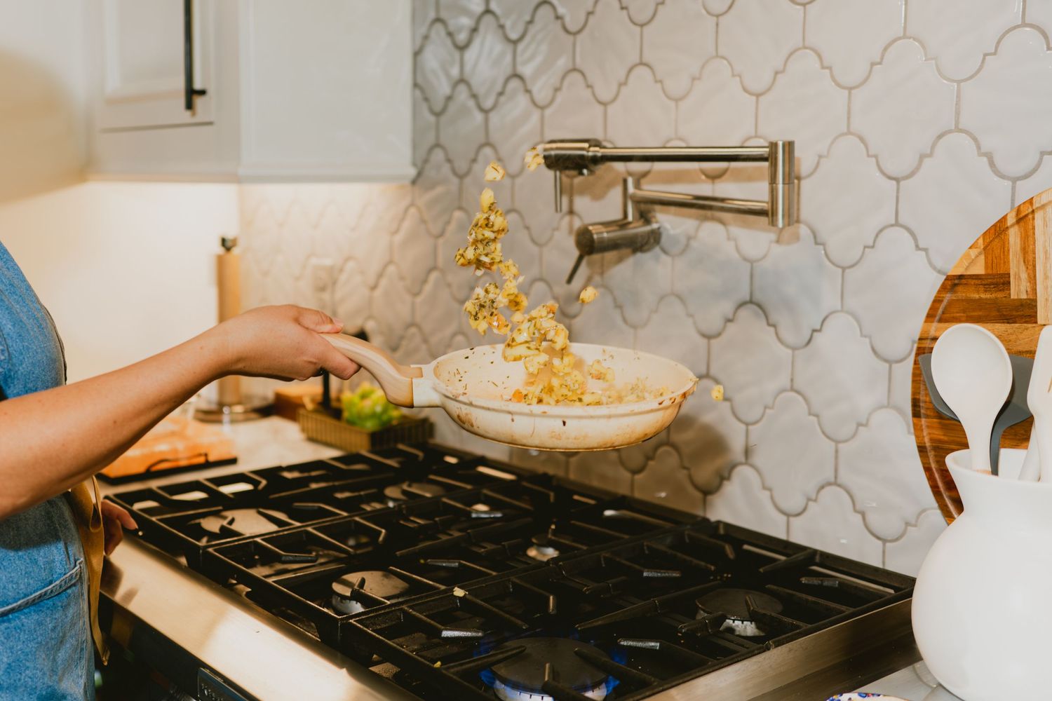 Person tossing ingredients in a bowl over a gas stovetop with a decorative tile backsplash in a modern kitchen.
