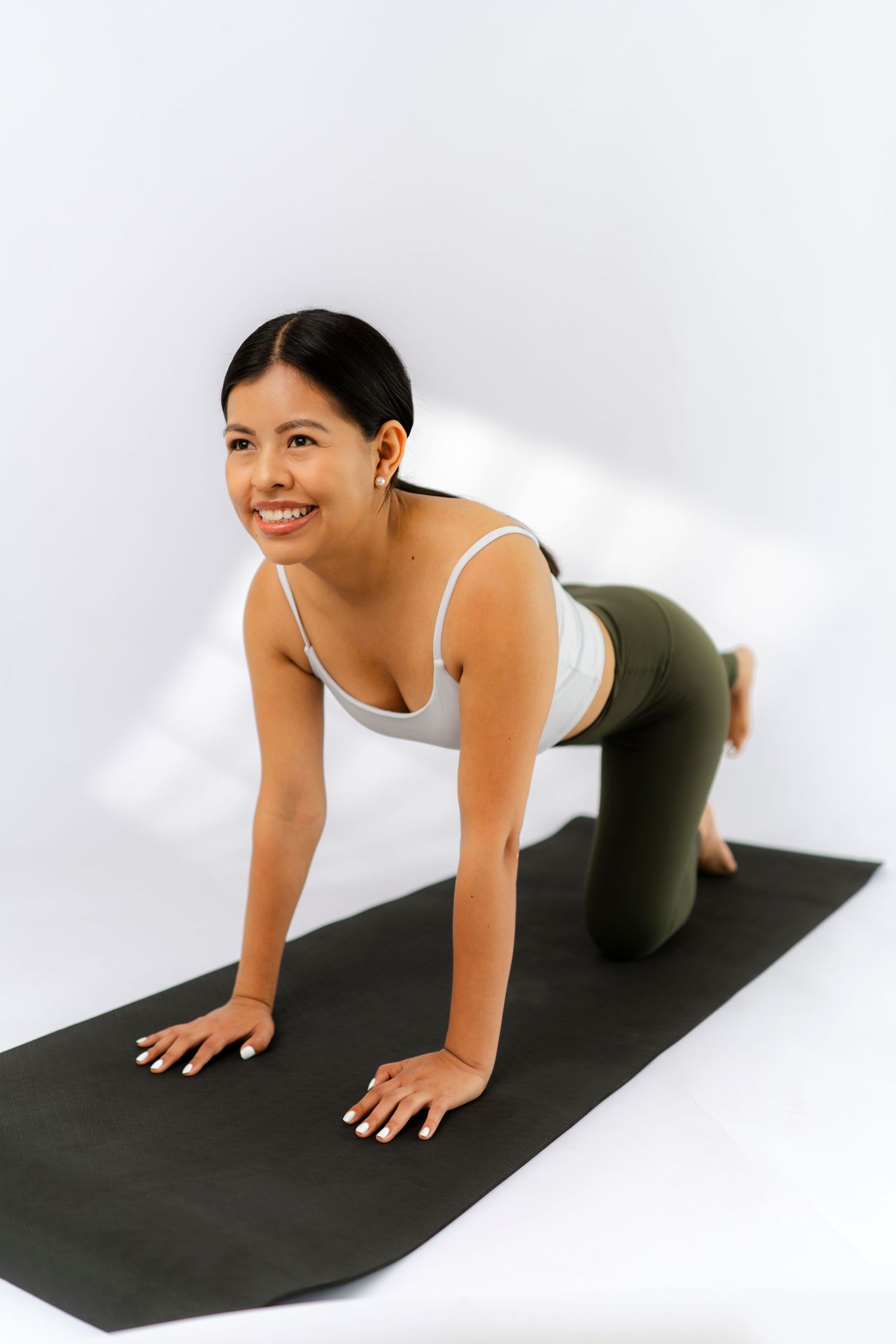 Smiling woman in white sports bra and green leggings doing yoga on a black mat against a white background.