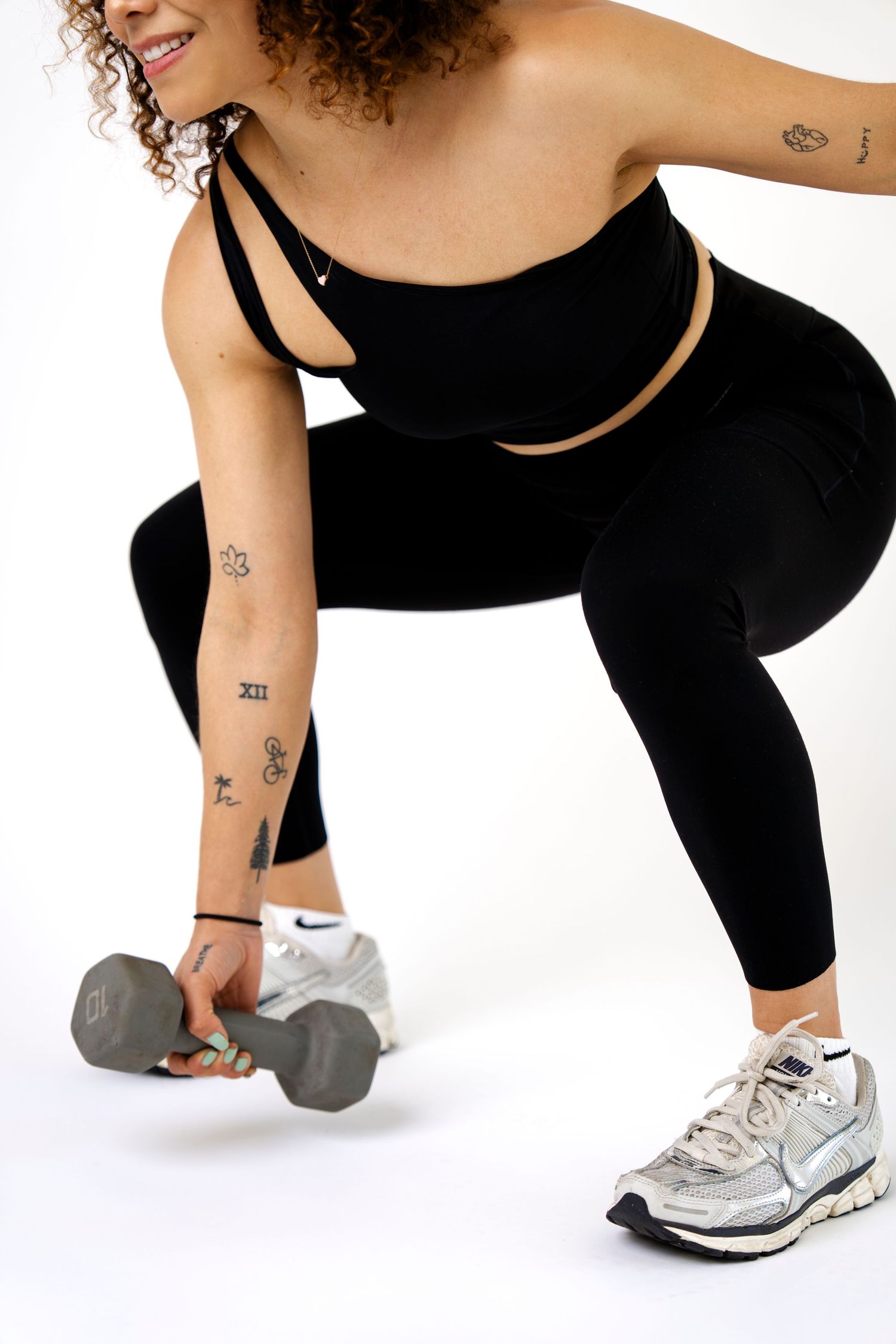Woman in black Nike sports bra and leggings doing a lunge while holding a dumbbell, with tattoos visible on her arm.