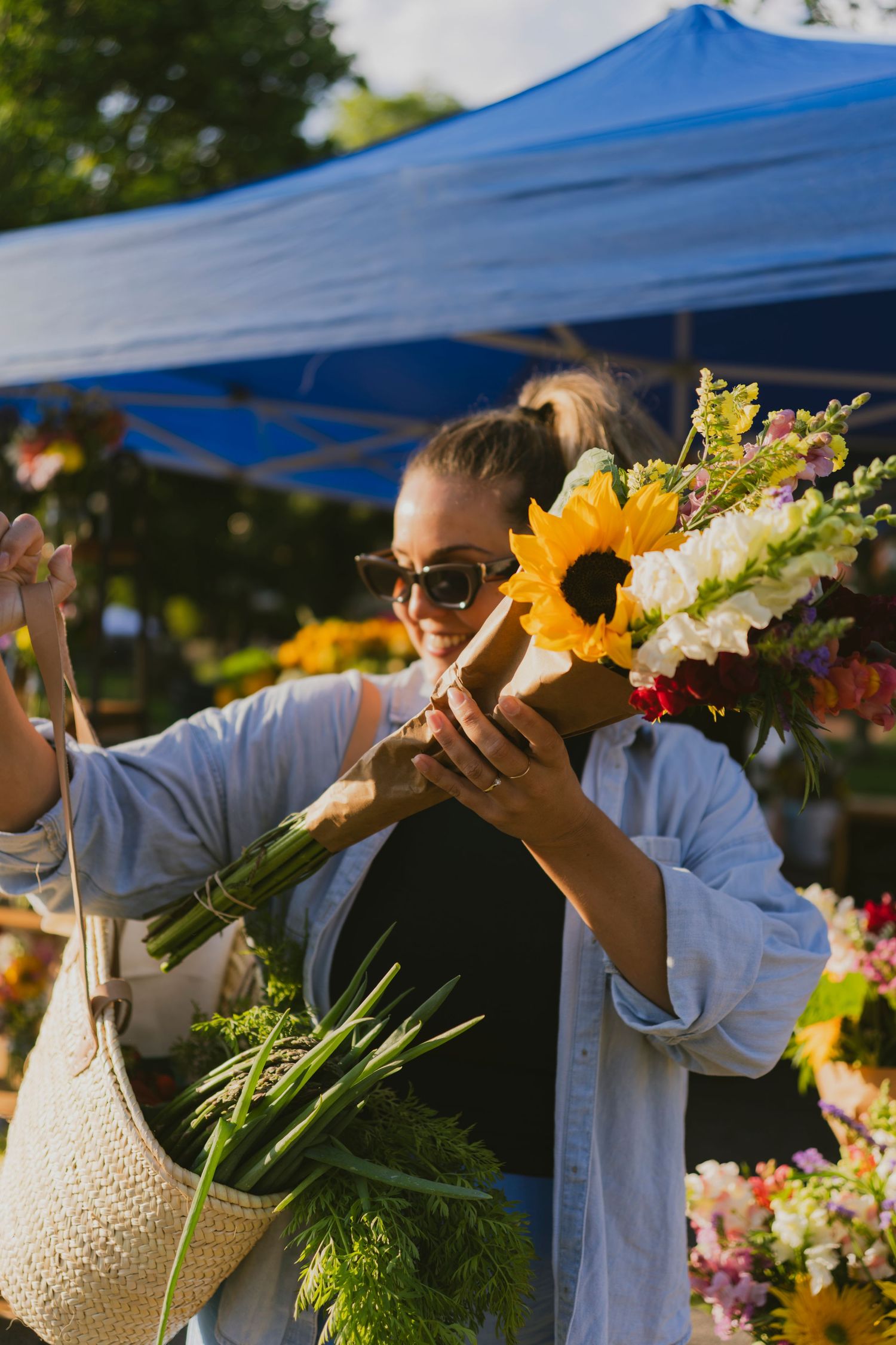 Woman in sunglasses smelling a sunflower bouquet at an outdoor farmers market flower stall on a sunny day.
