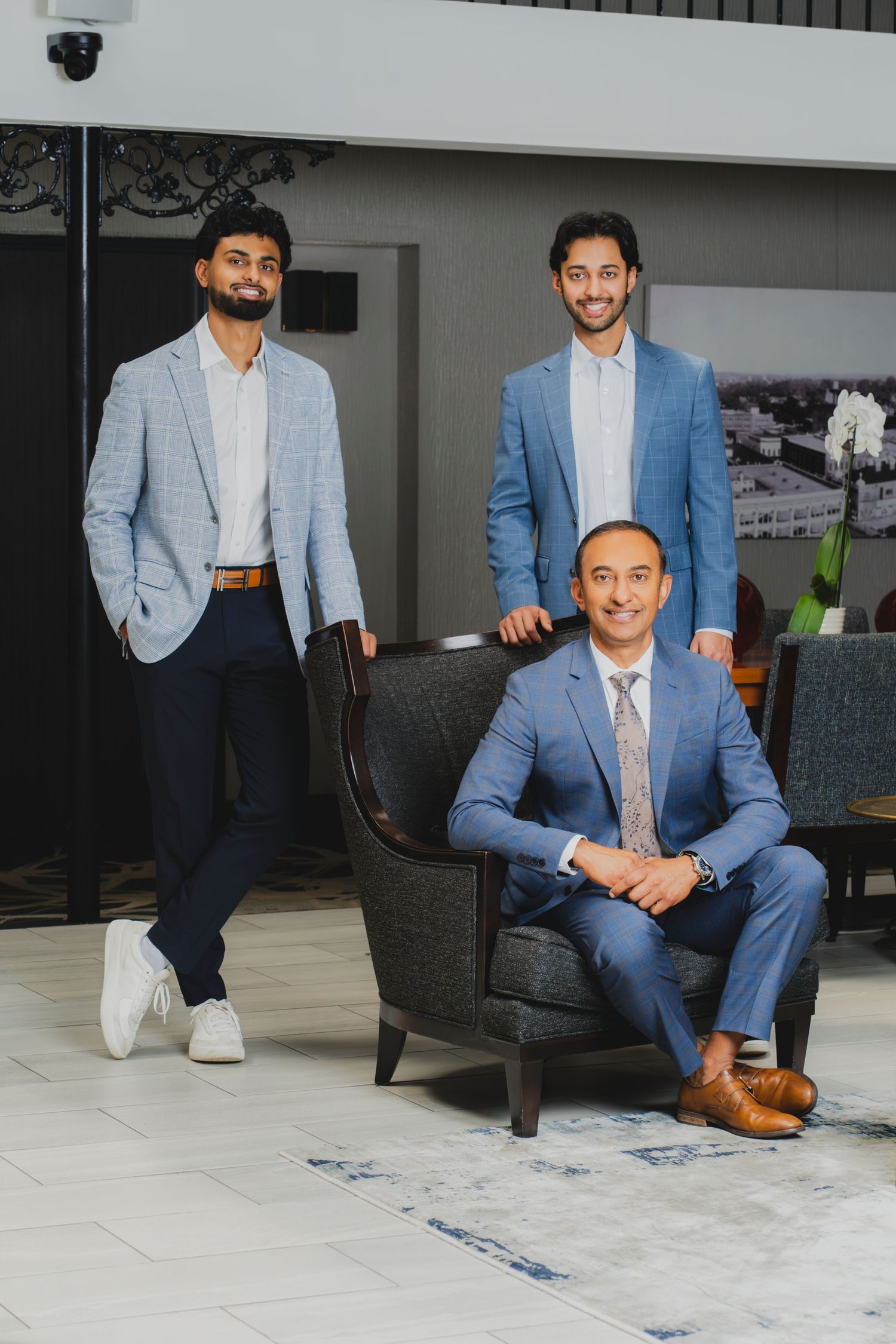 Three well-dressed men in blue suits pose professionally in a modern lobby, two standing and one seated in an armchair.