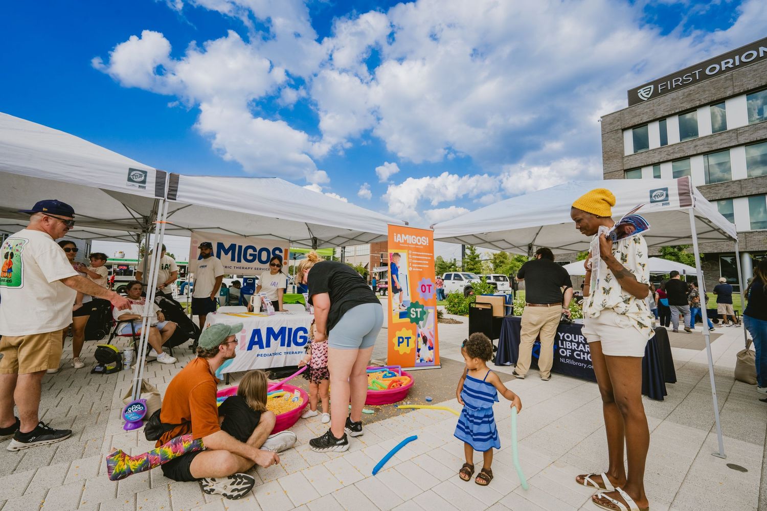Wide angle view of an outdoor community event with tents and activities under puffy clouds.