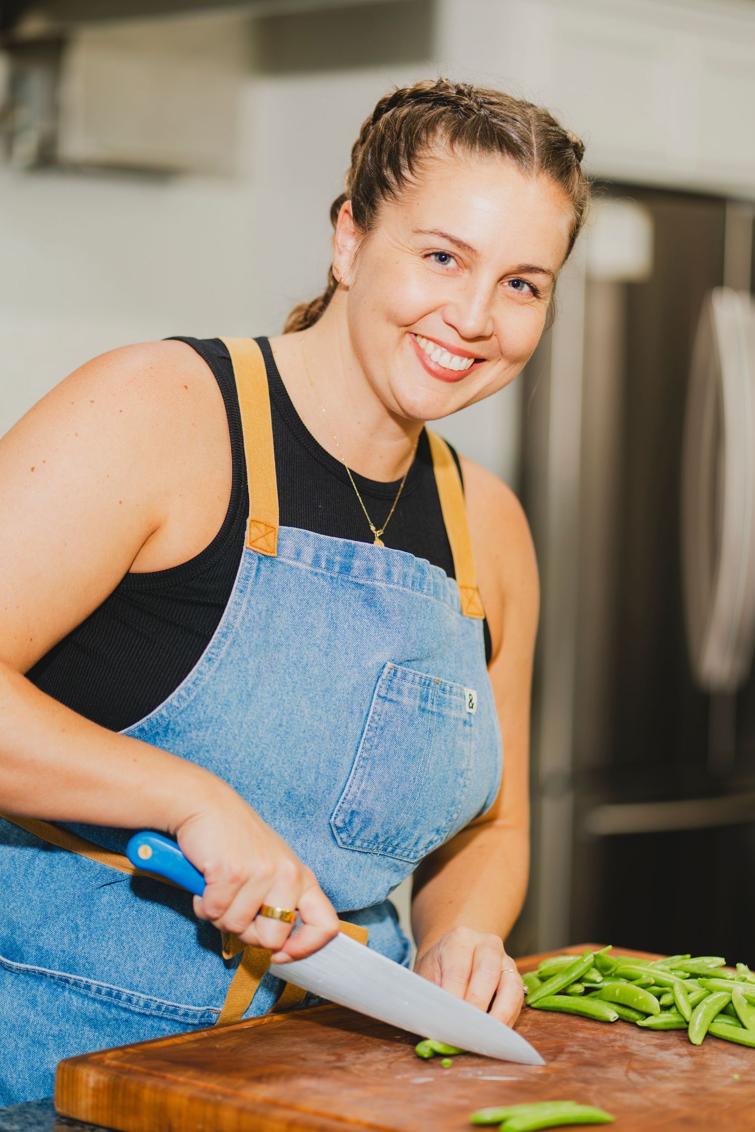Smiling woman in denim apron chopping fresh green beans on wooden cutting board in modern kitchen.