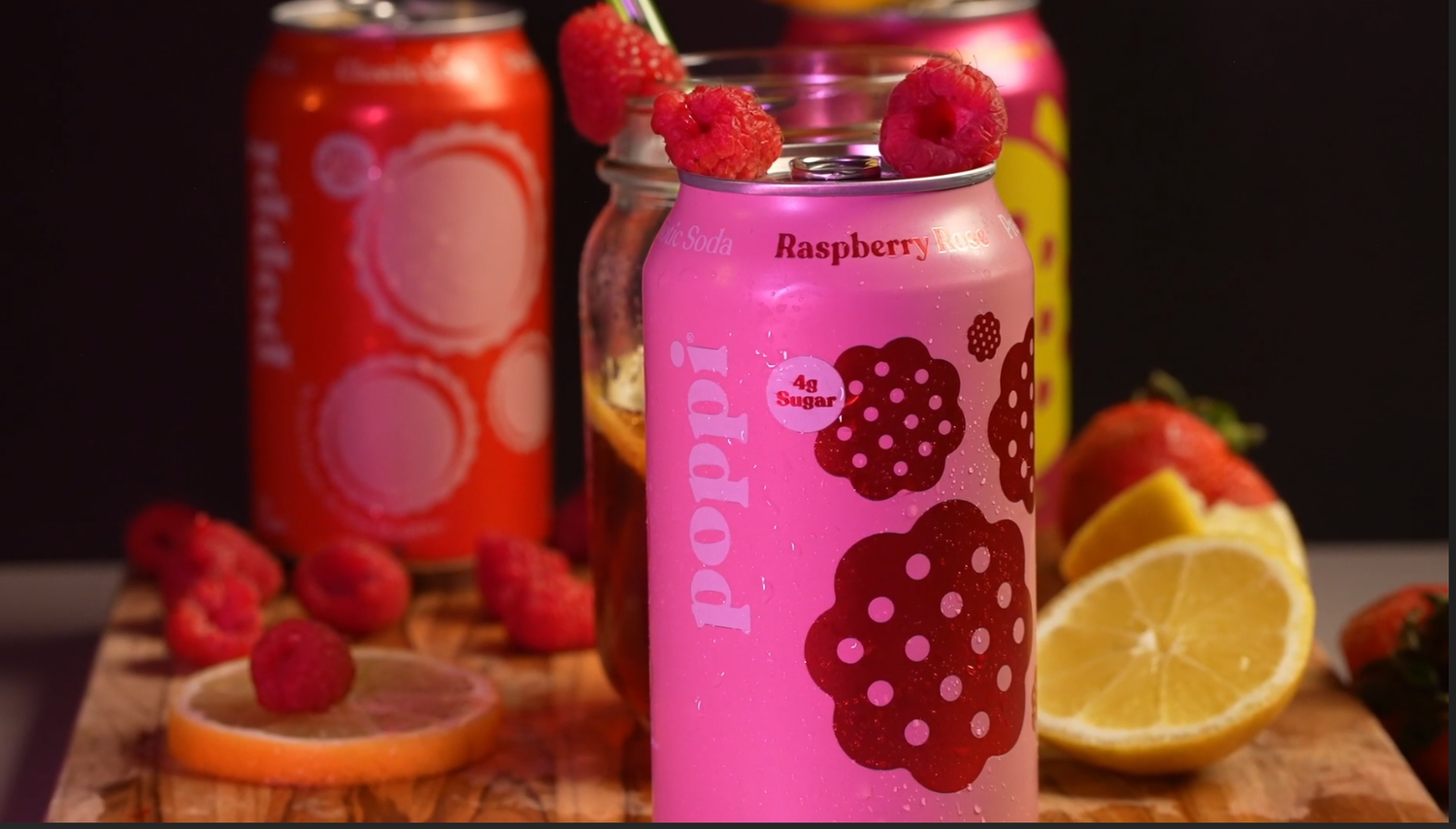 Bright pink canned beverages with raspberry branding arranged with fresh fruit. poppi
