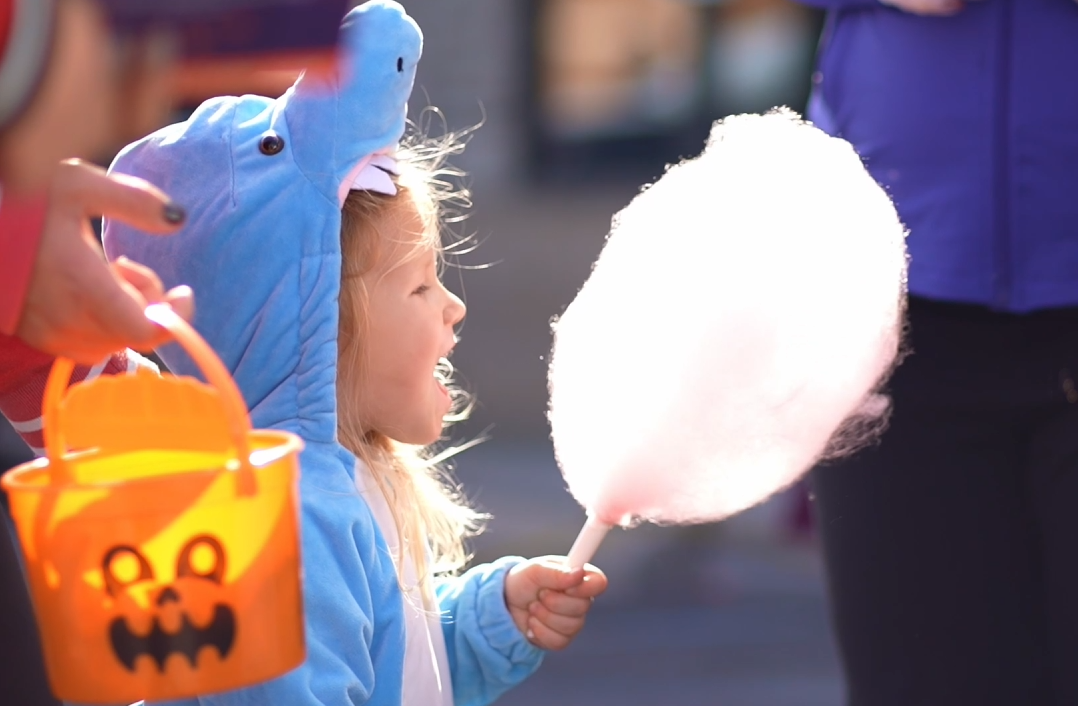 Child in blue costume holding cotton candy and orange Halloween bucket at festival. Trunk or treat. Halloween. Trick or treat.