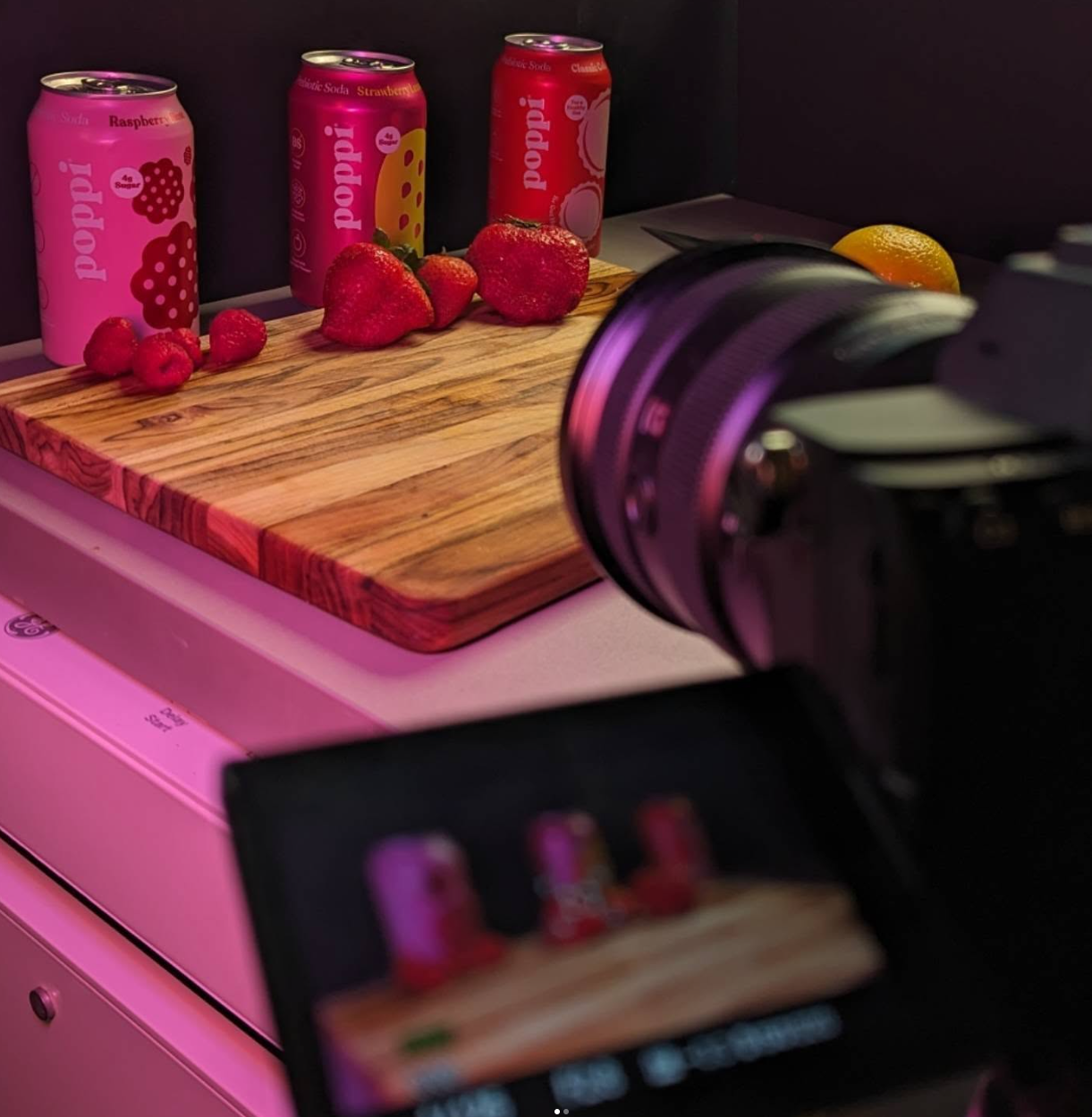 Pink canned beverages on wooden cutting board with camera lens in foreground. product videography. product photography. drink poppi