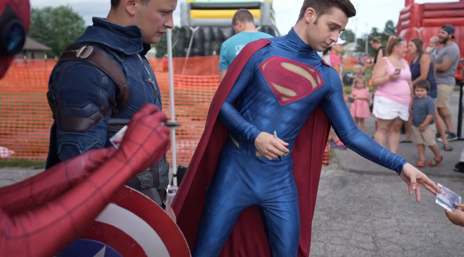 People in Superman and Captain America costumes at an outdoor event, interacting with fans at a cosplay gathering.