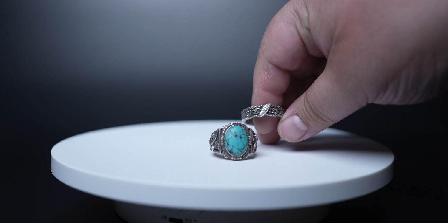 Hand picking up a silver ring with turquoise stone on a white display stand against dark background.