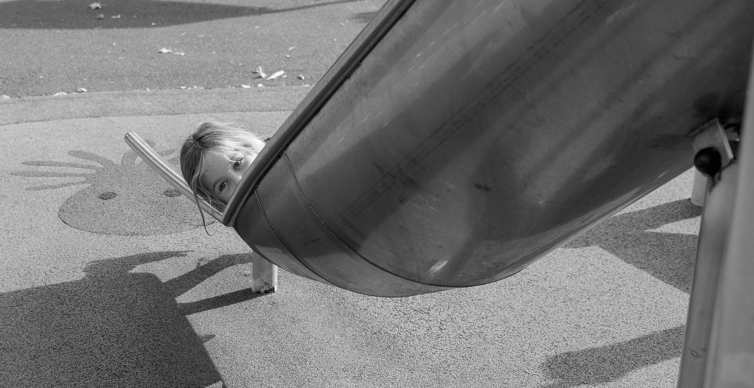A black and white photo shows someone playfully peeking out from behind a slide at a playground.