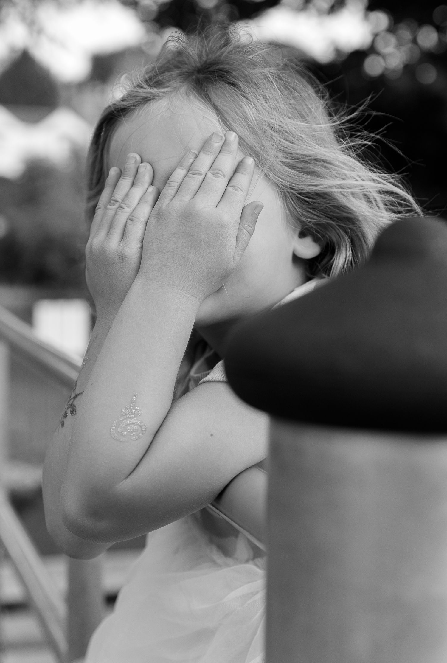 Black and white portrait of a child covering their face with both hands outdoors.
