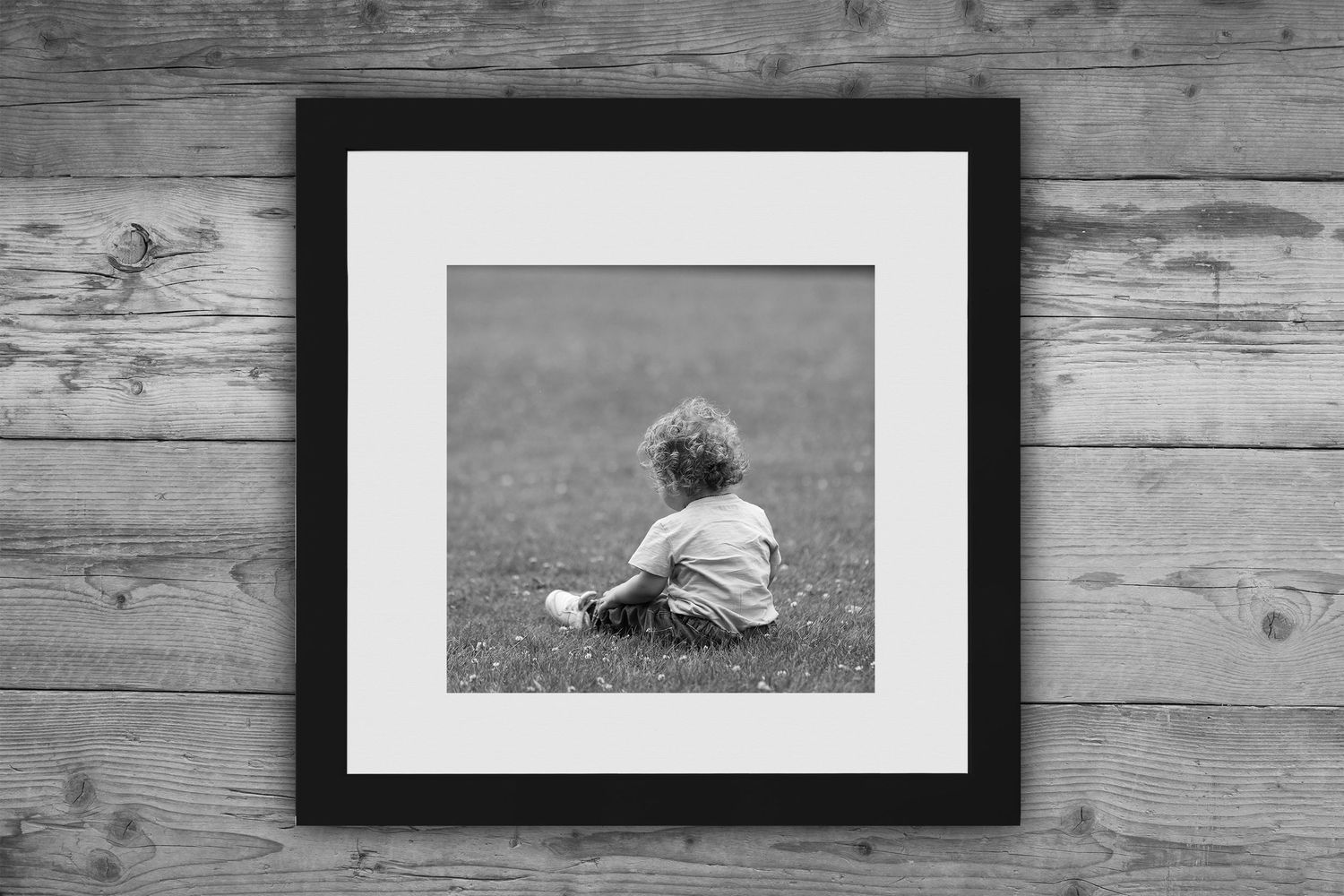 Black and white framed photograph of a small child sitting alone on grass, displayed on rustic wooden wall.