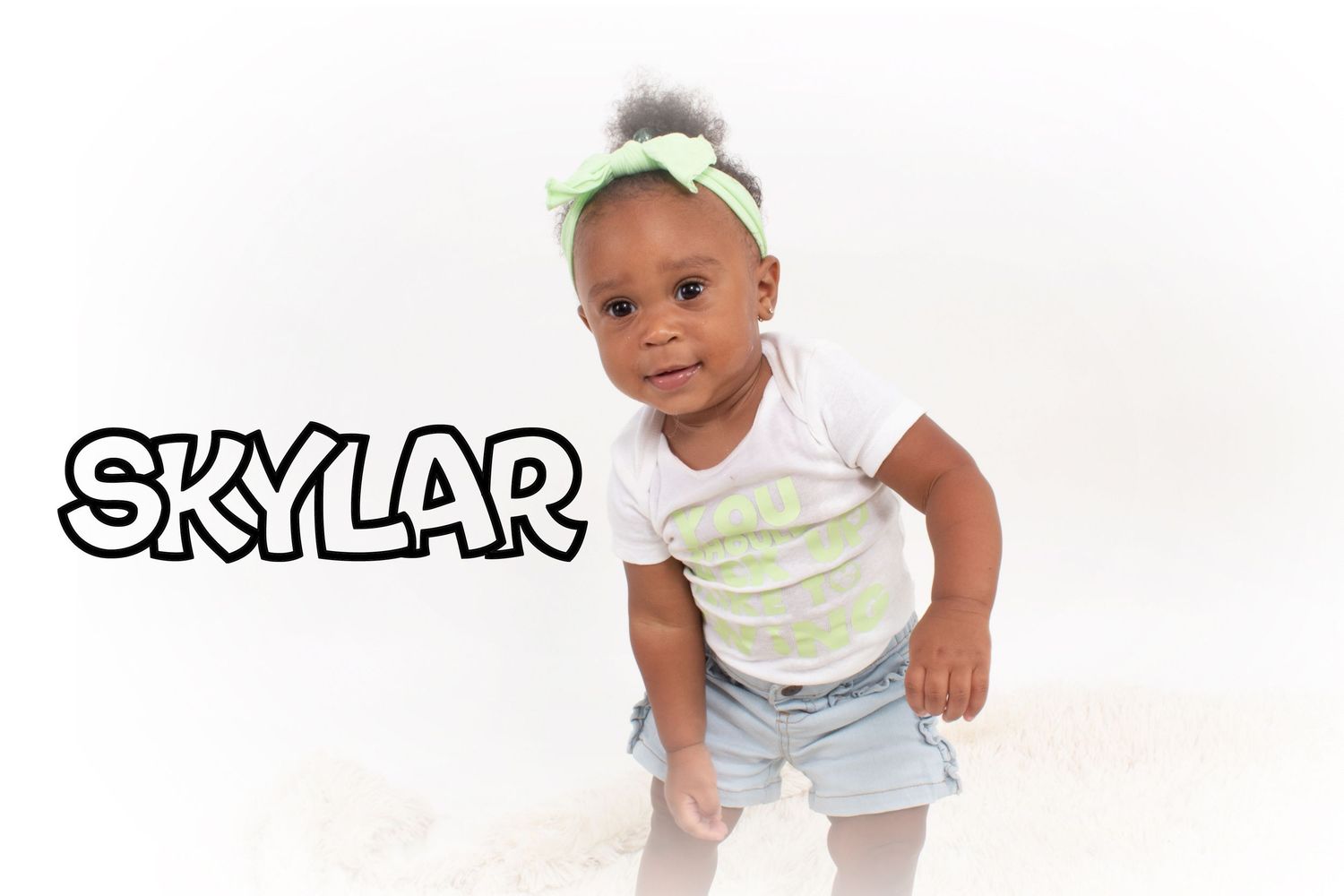 A toddler named Skylar poses playfully in a white shirt and denim skirt with a green headband.