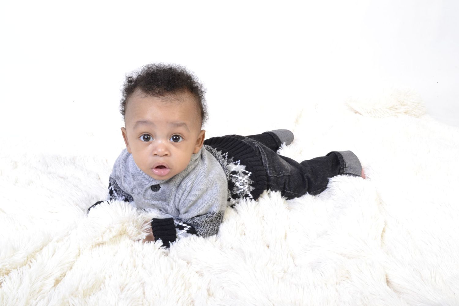 Adorable baby in gray and black outfit lies on fluffy white blanket looking at camera.