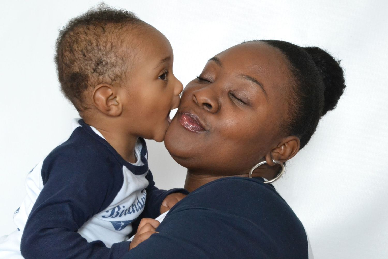 Adorable toddler gives his smiling mother a sweet kiss on the cheek in a heartwarming studio portrait.