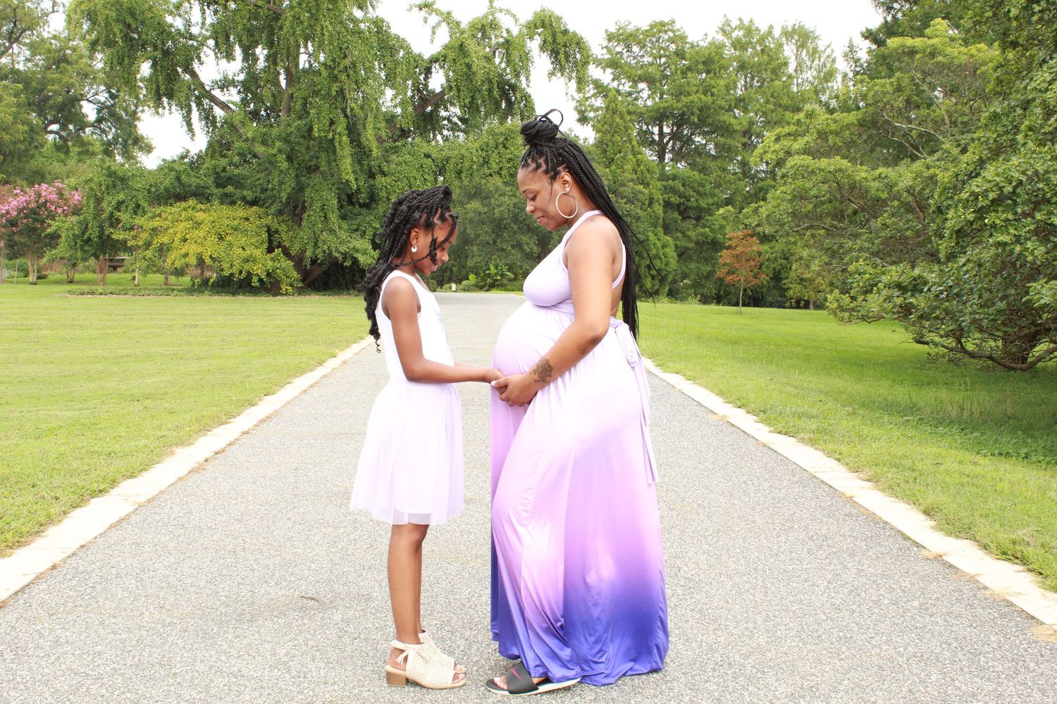 A pregnant woman in a purple ombre gown holds hands with a young girl in white on a park pathway.