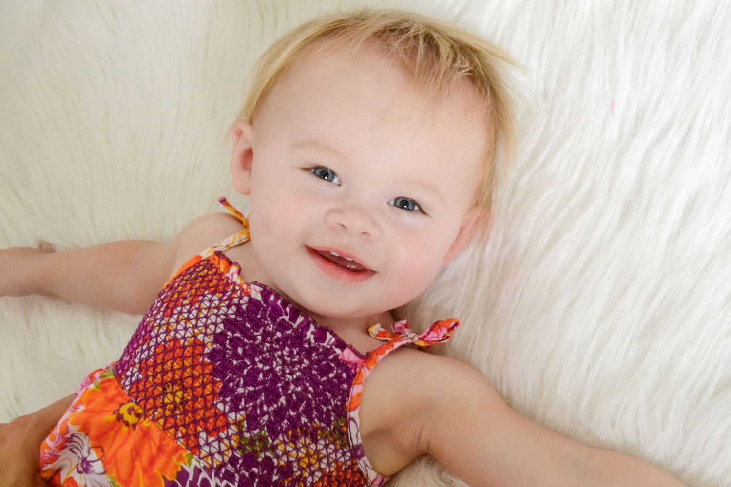 Smiling baby girl in colorful floral romper lying on white fluffy blanket, looking up with bright blue eyes.
