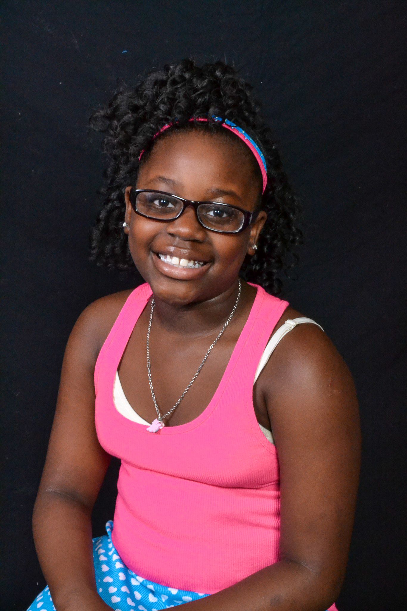 Smiling young girl with curly hair, glasses, and pink tank top poses for a portrait against a dark background.