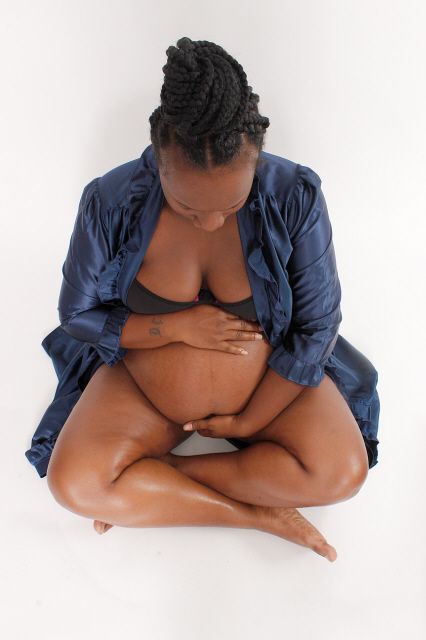 Pregnant Black woman in navy robe and black bikini top sits cross-legged on floor cradling her belly.