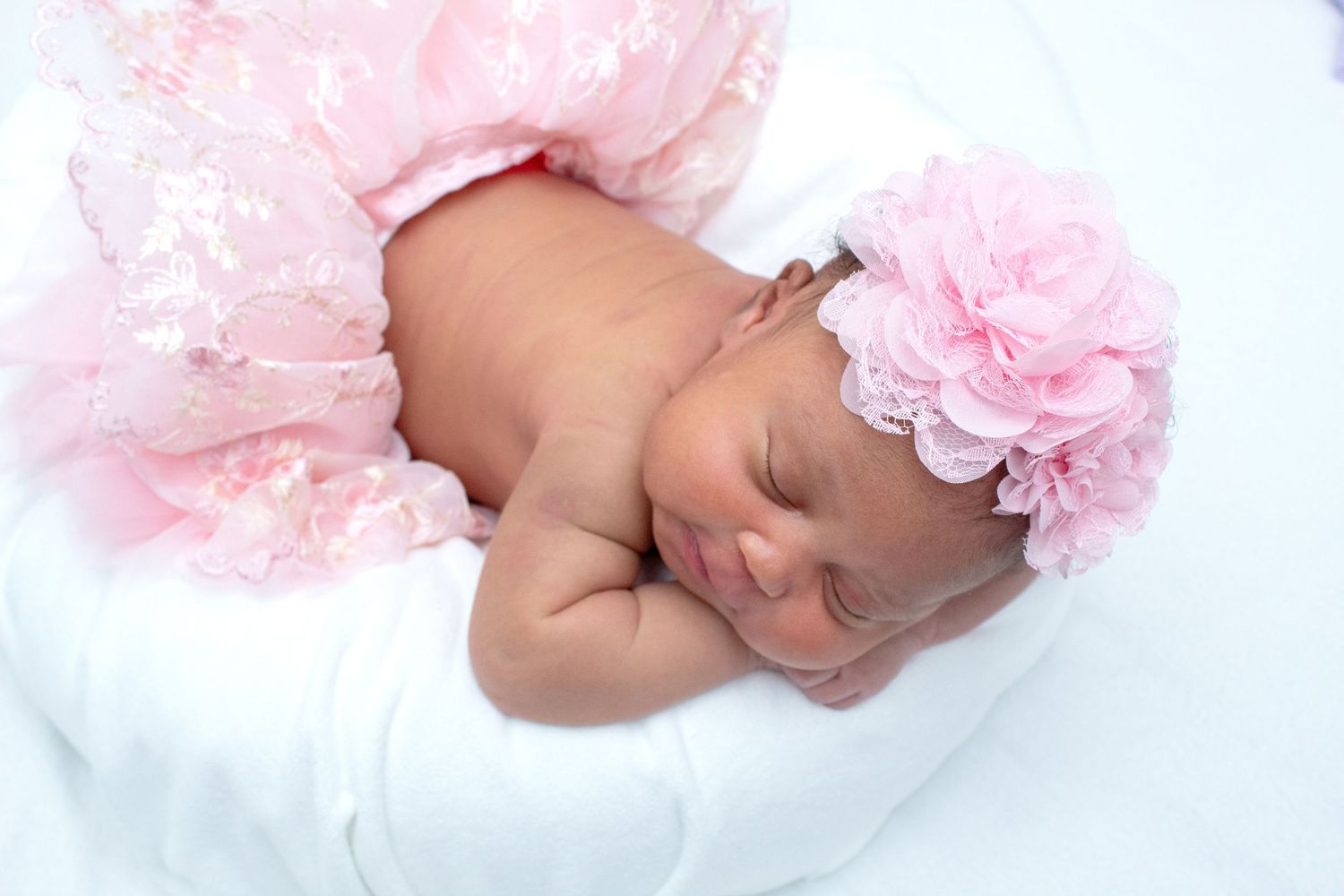 Newborn baby girl in pink lace outfit and floral headband sleeping on white pillow.