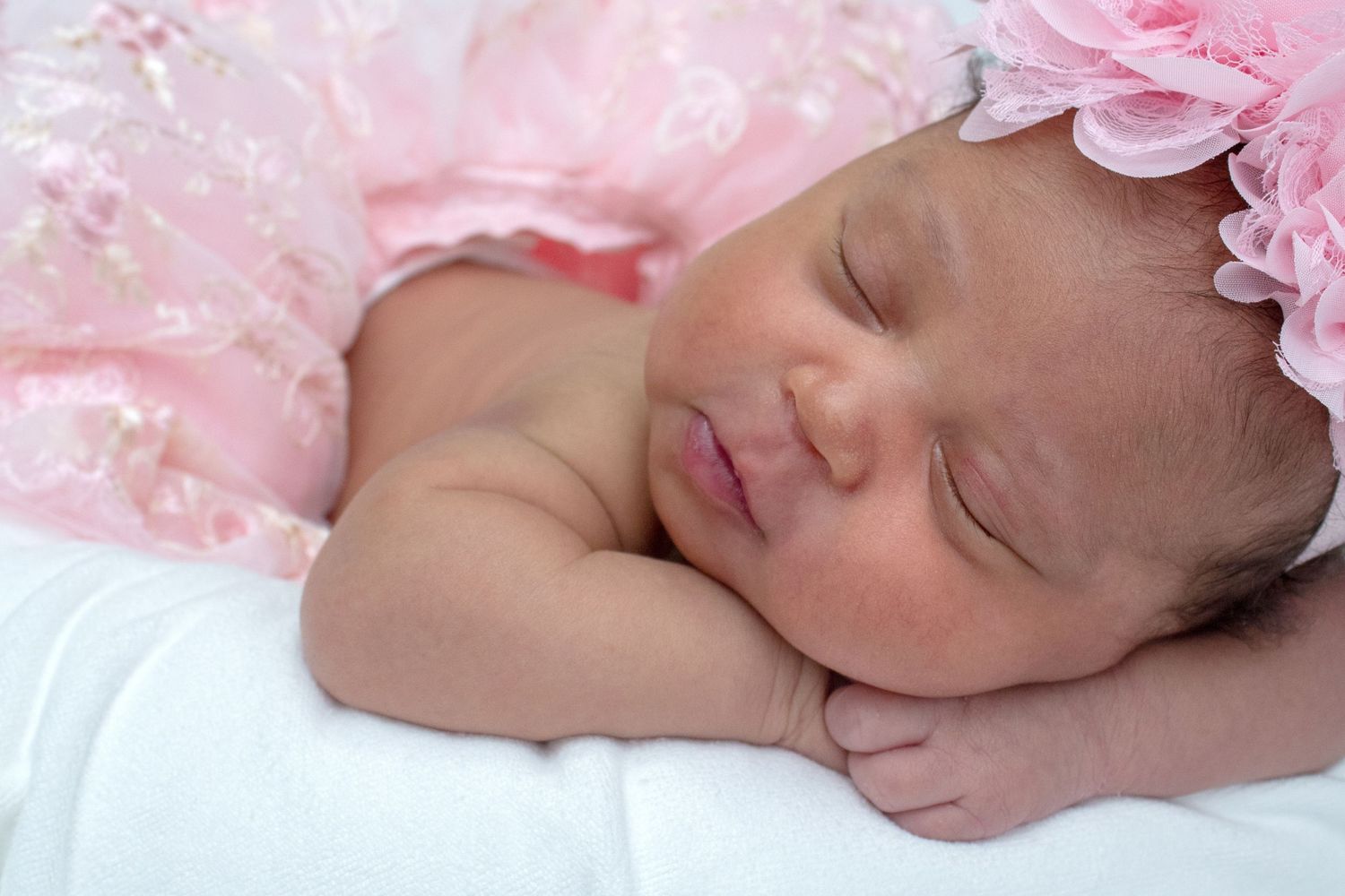 Peaceful newborn baby girl sleeping in pink dress and floral headband on white blanket.
