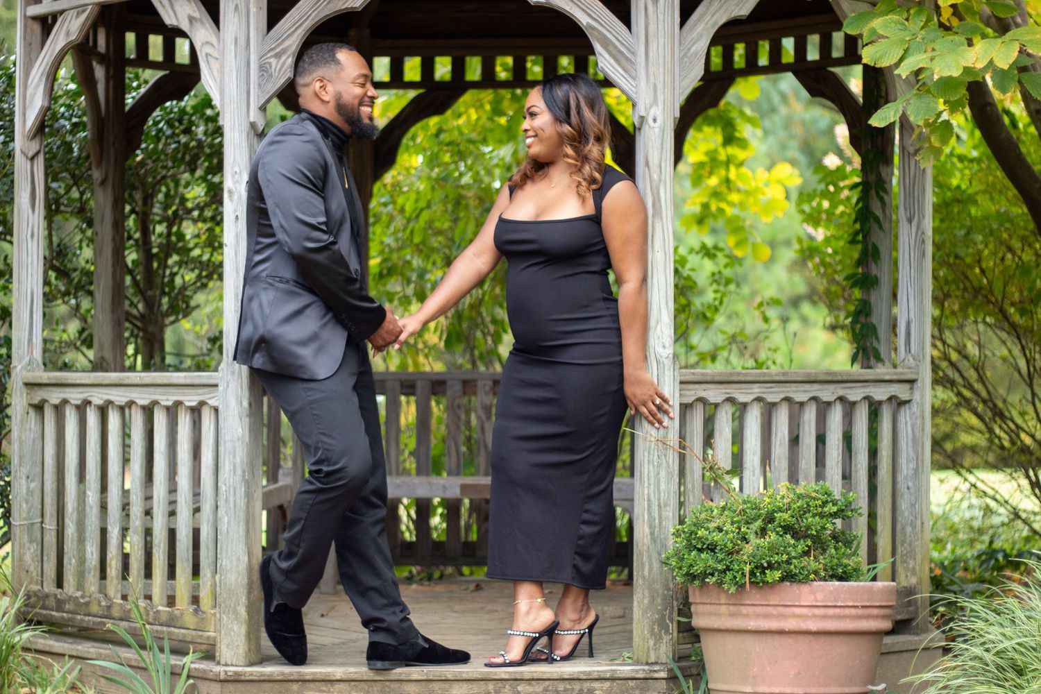Couple in black attire holding hands under a wooden gazebo surrounded by lush greenery.