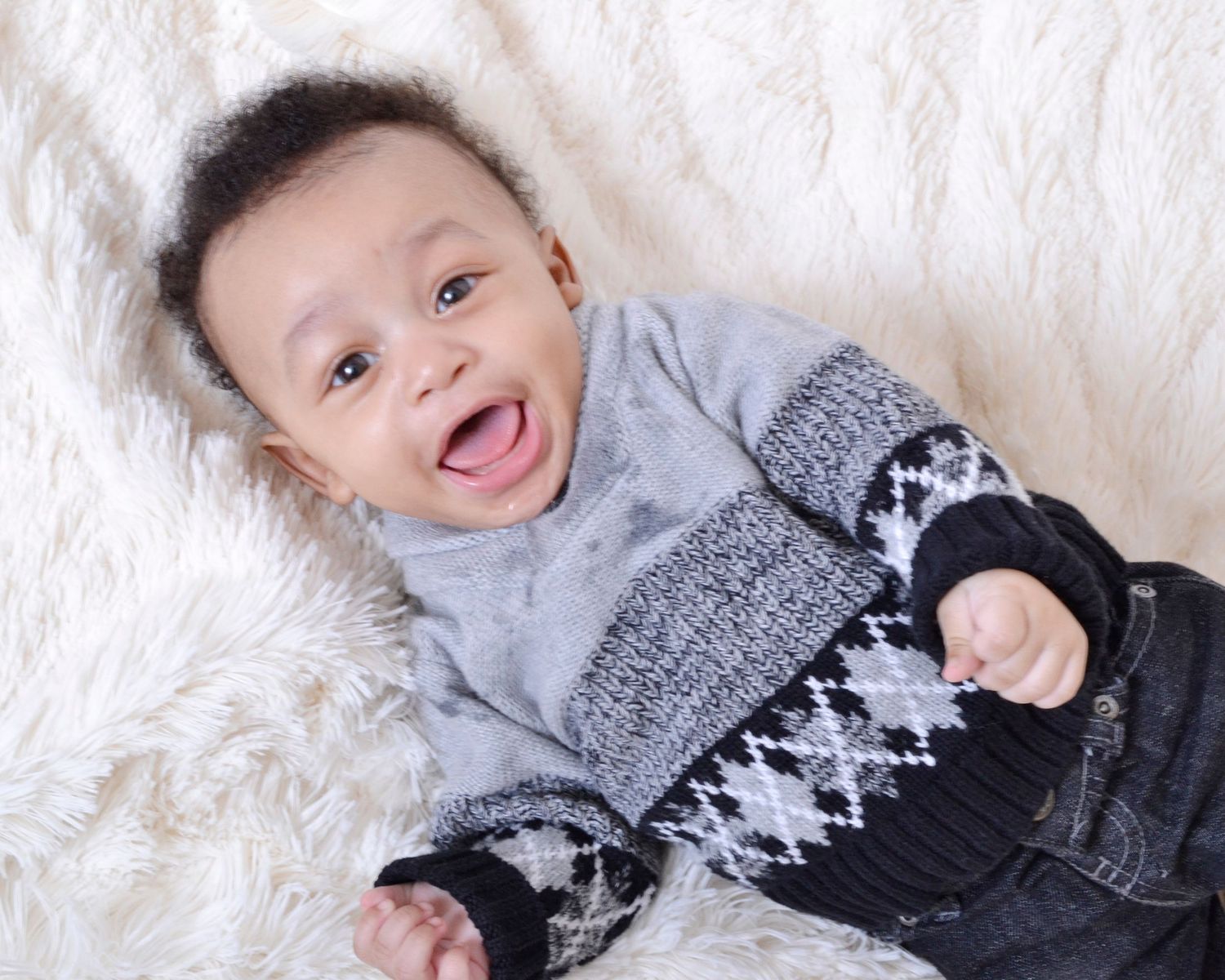 Smiling baby in gray and black knit sweater lying on white fluffy blanket, looking up with joy.