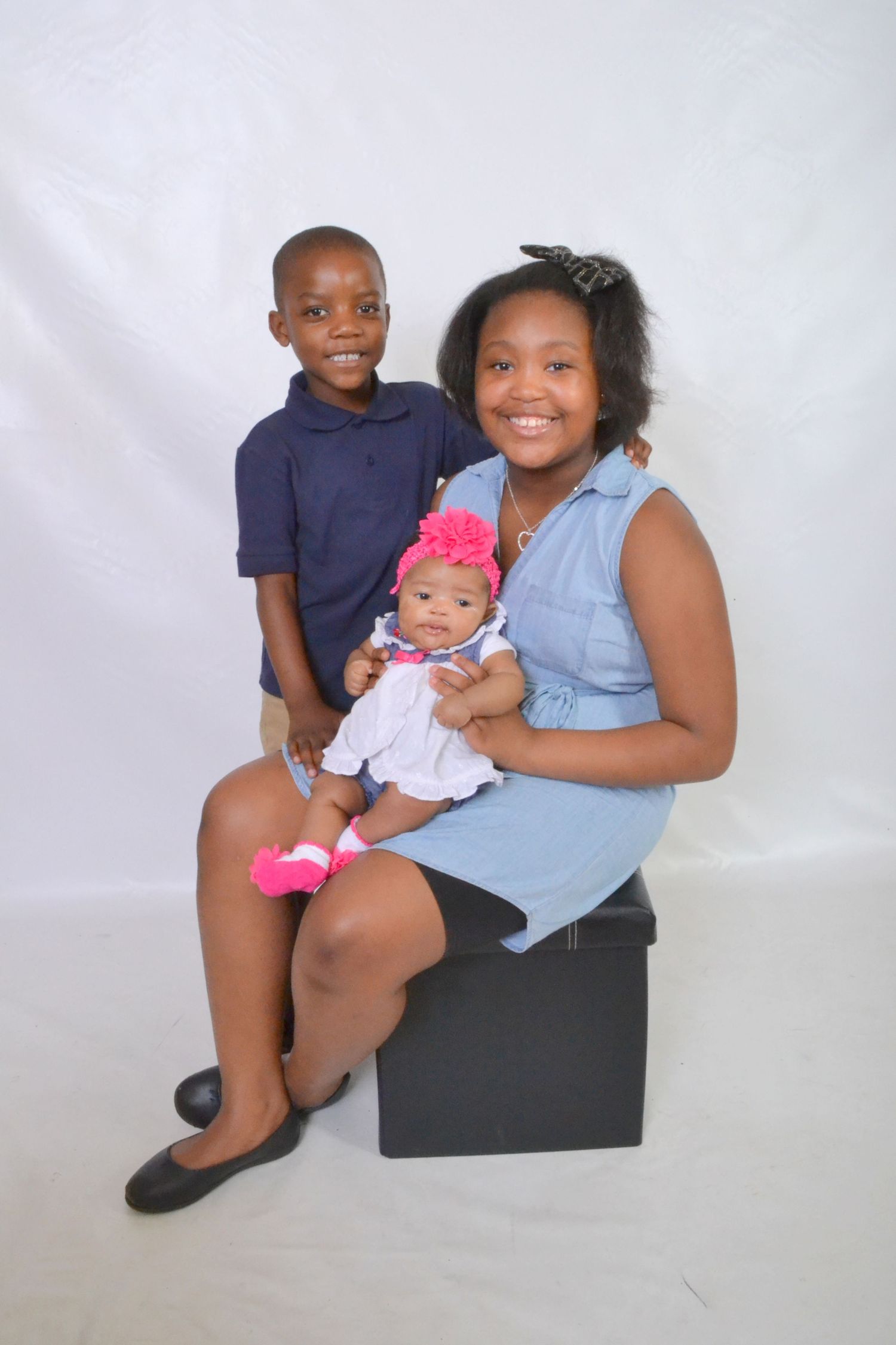 Young girl sitting on black box holding infant with boy standing beside her on white background.