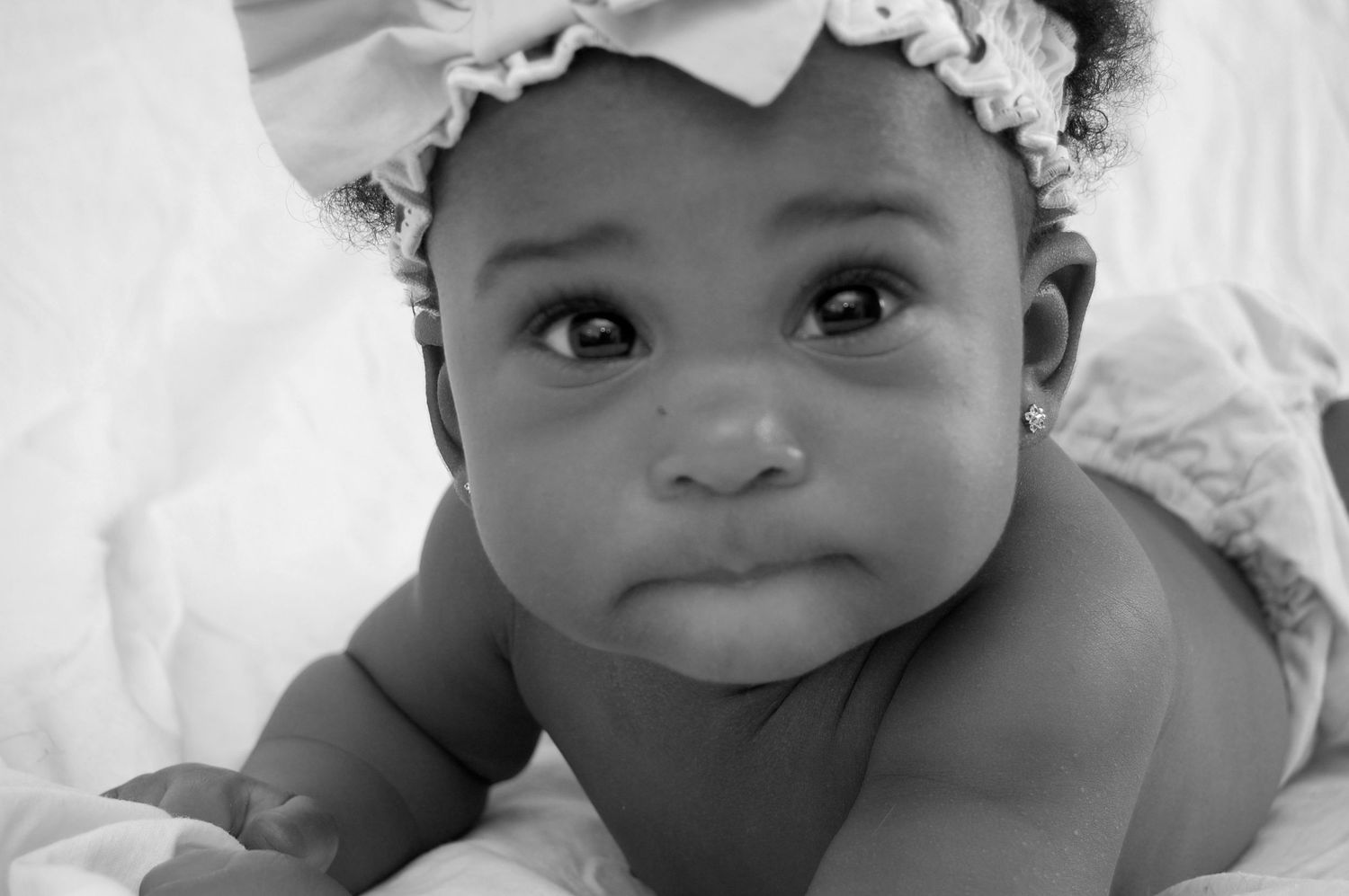 Black and white portrait of an adorable baby girl wearing a floral headband, looking up with big expressive eyes.