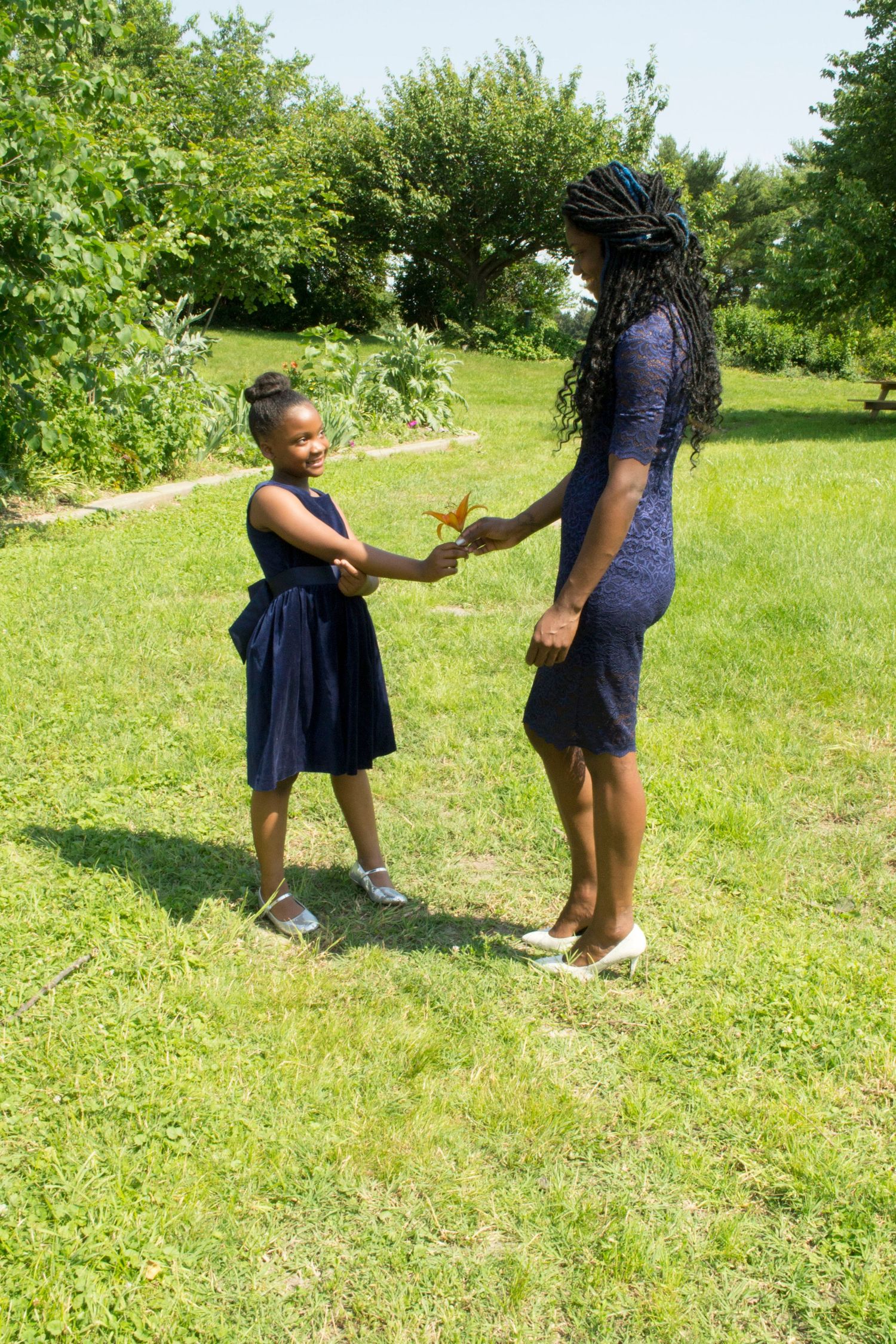 A woman and young girl in navy blue outfits share a flower while holding hands in a sunny green park.