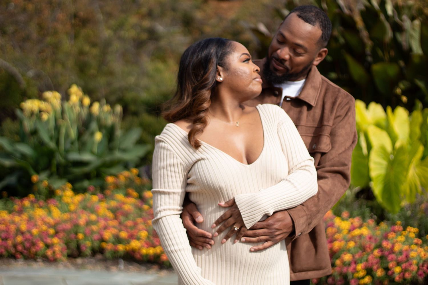 Expecting couple embracing outdoors in garden with colorful flowers during maternity photoshoot.