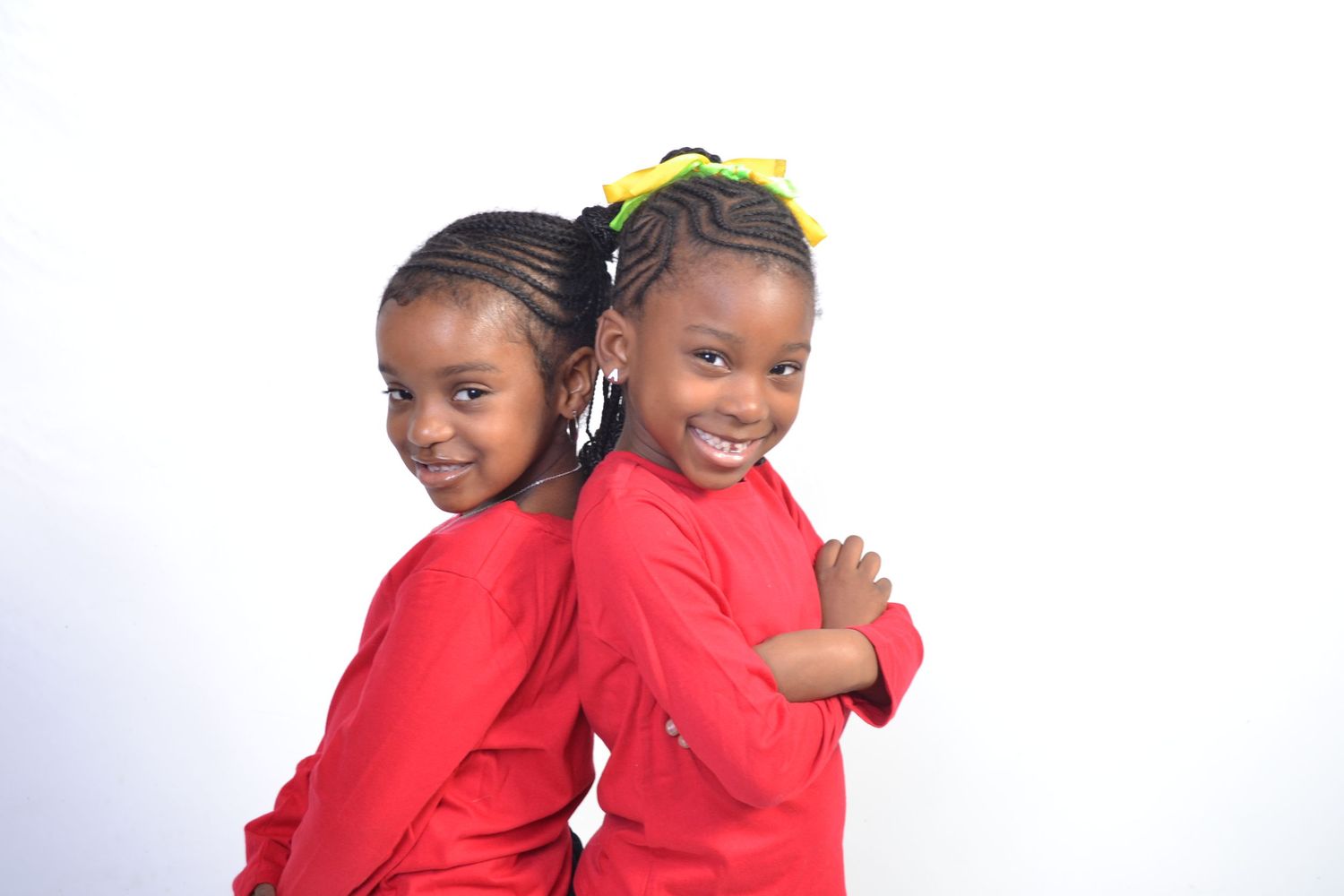 Two young girls in red shirts pose back to back smiling on white background.