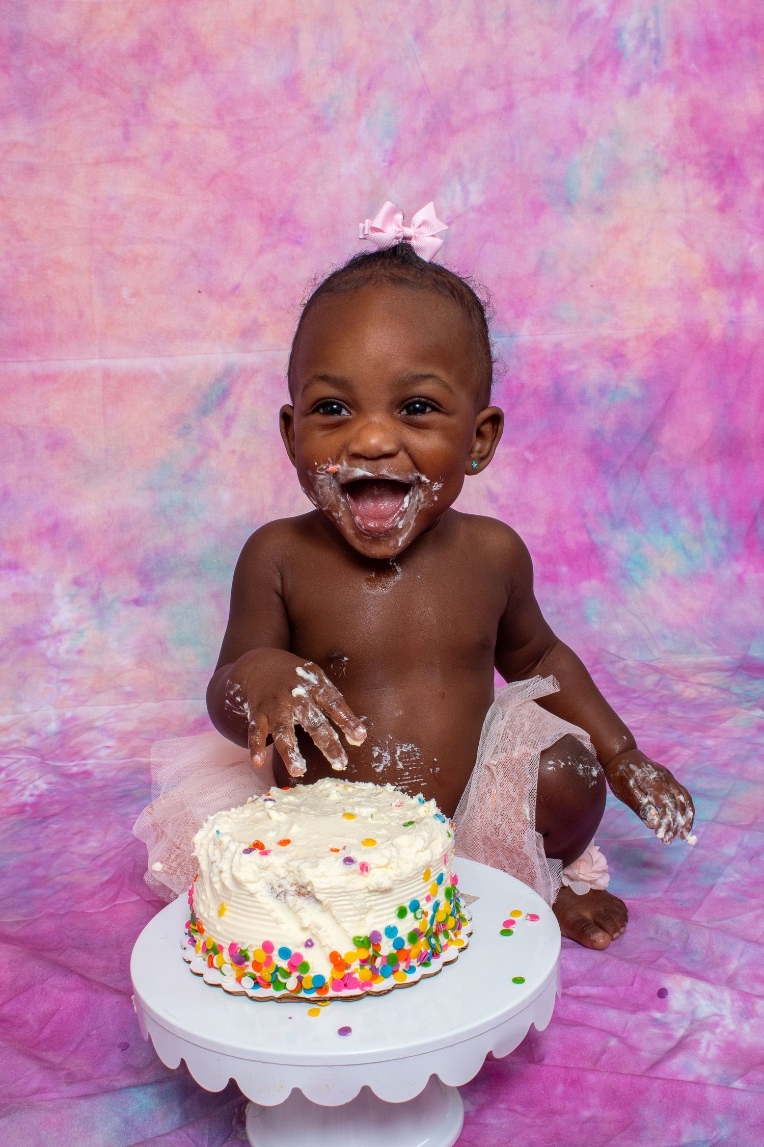 Joyful baby girl in pink tutu smashes first birthday cake, covered in white frosting, against colorful backdrop.
