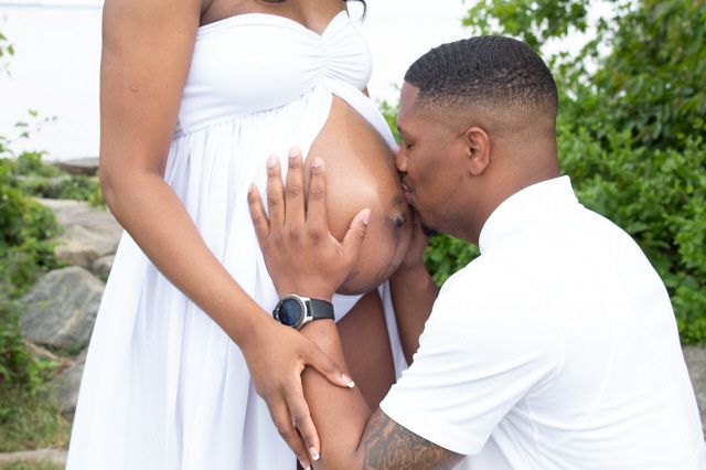 Man in white shirt kissing pregnant woman's belly during outdoor maternity photoshoot.