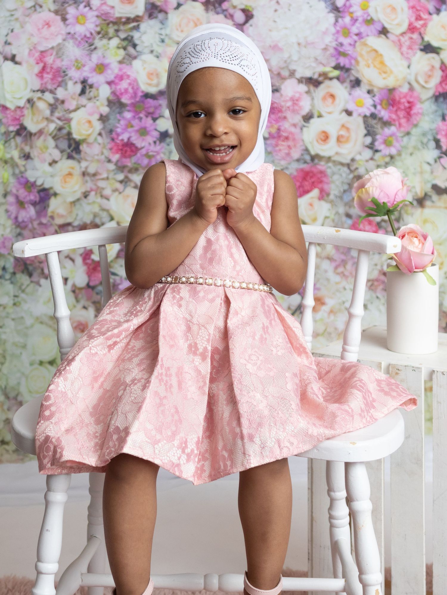 Toddler girl in pink floral dress and white hijab smiling in front of flower wall backdrop.