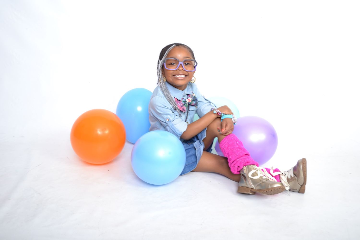 Young girl smiling with colorful balloons on white background, wearing glasses and denim jacket.