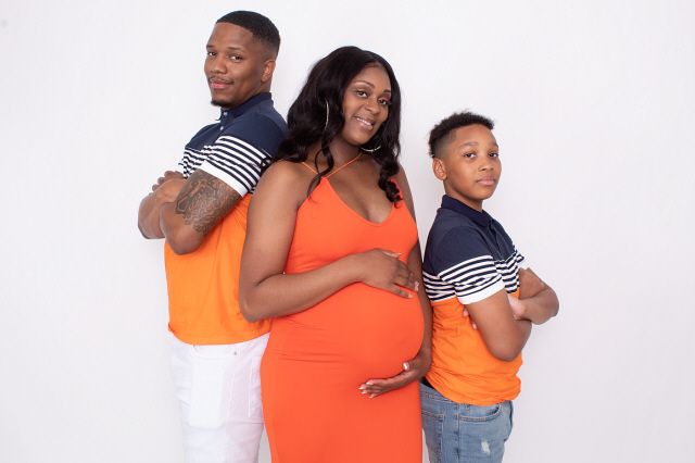 A pregnant woman in an orange dress poses with two males in matching orange shirts for a maternity photo.