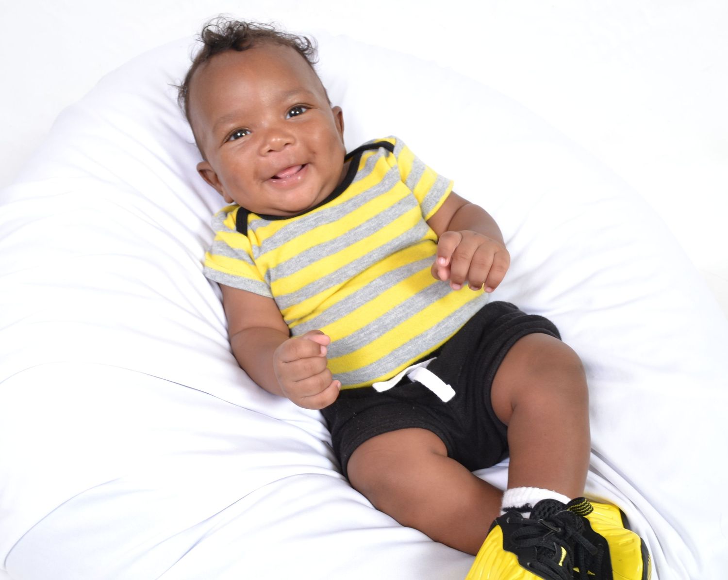 Happy baby boy in yellow striped onesie and black shorts sitting and smiling on white background.