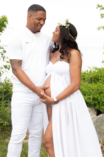 Pregnant couple in white outfits holding baby bump outdoors during maternity photoshoot.