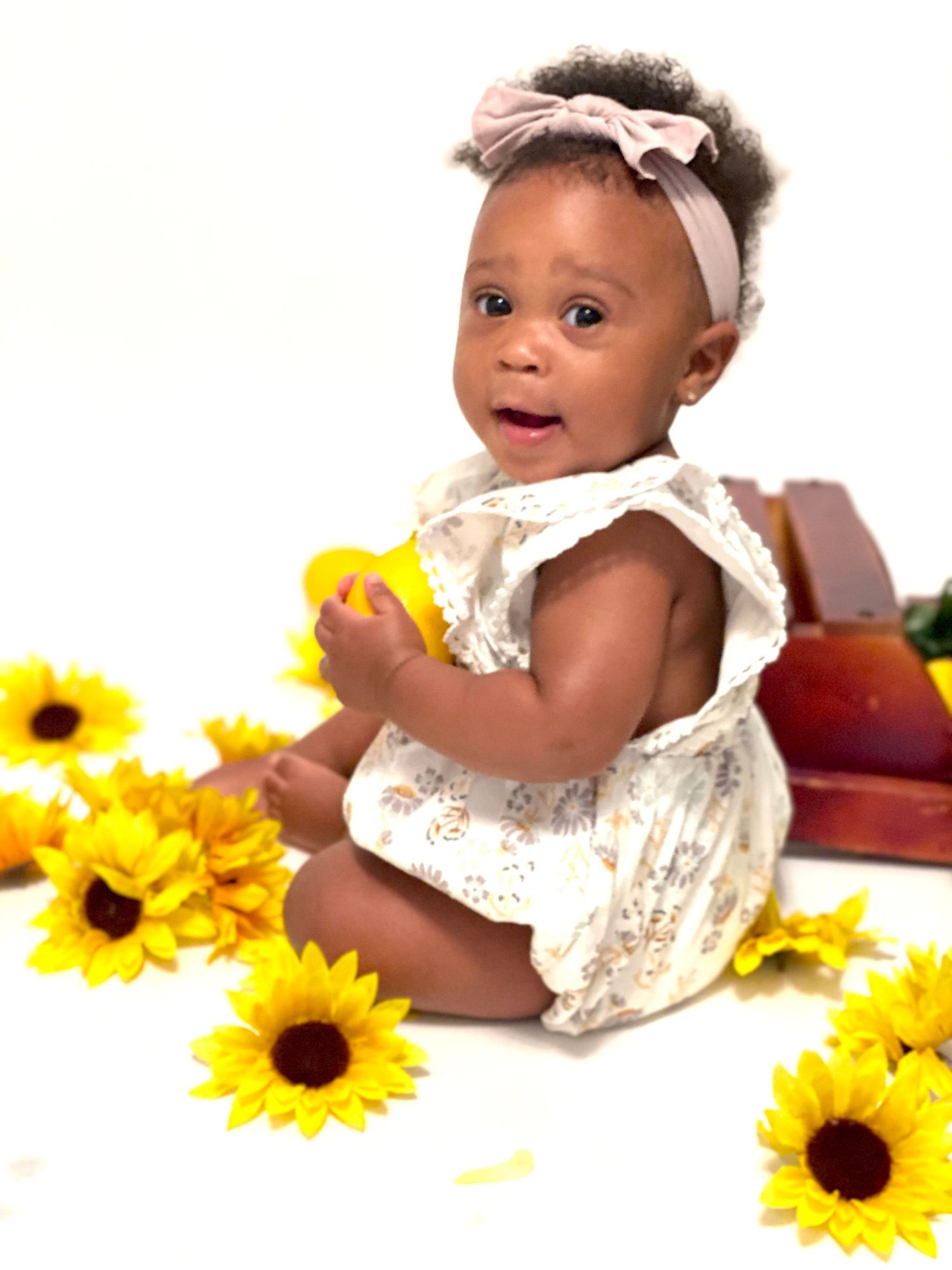 Adorable baby girl in floral romper and pink bow headband sitting among yellow sunflowers on white background.