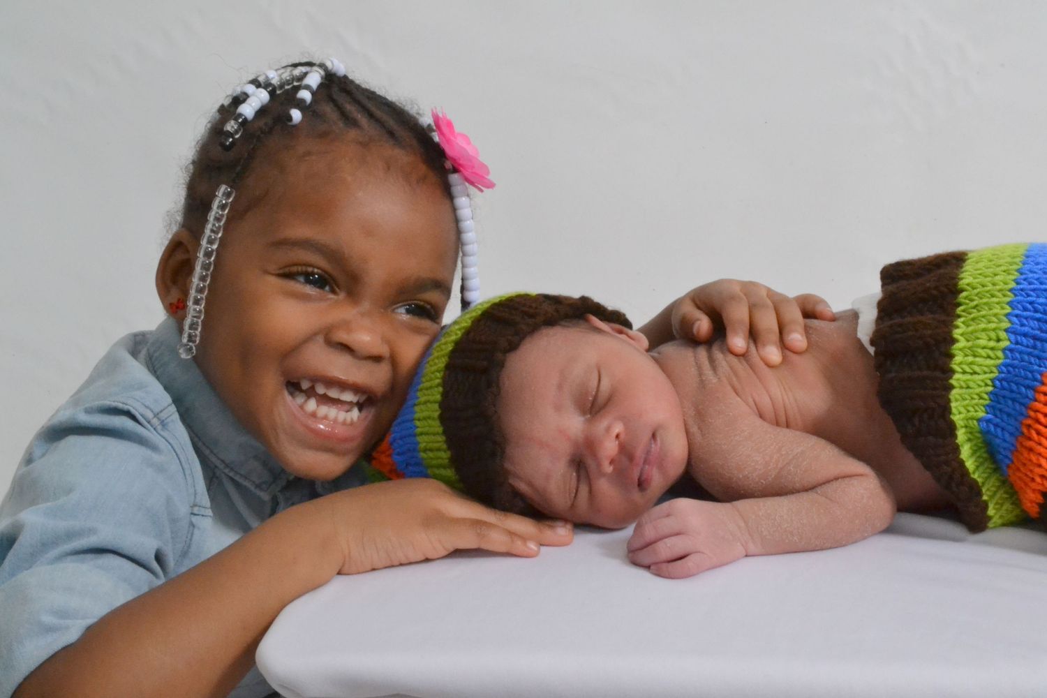 A smiling young girl with braids poses next to a sleeping newborn baby wrapped in a colorful knitted outfit.