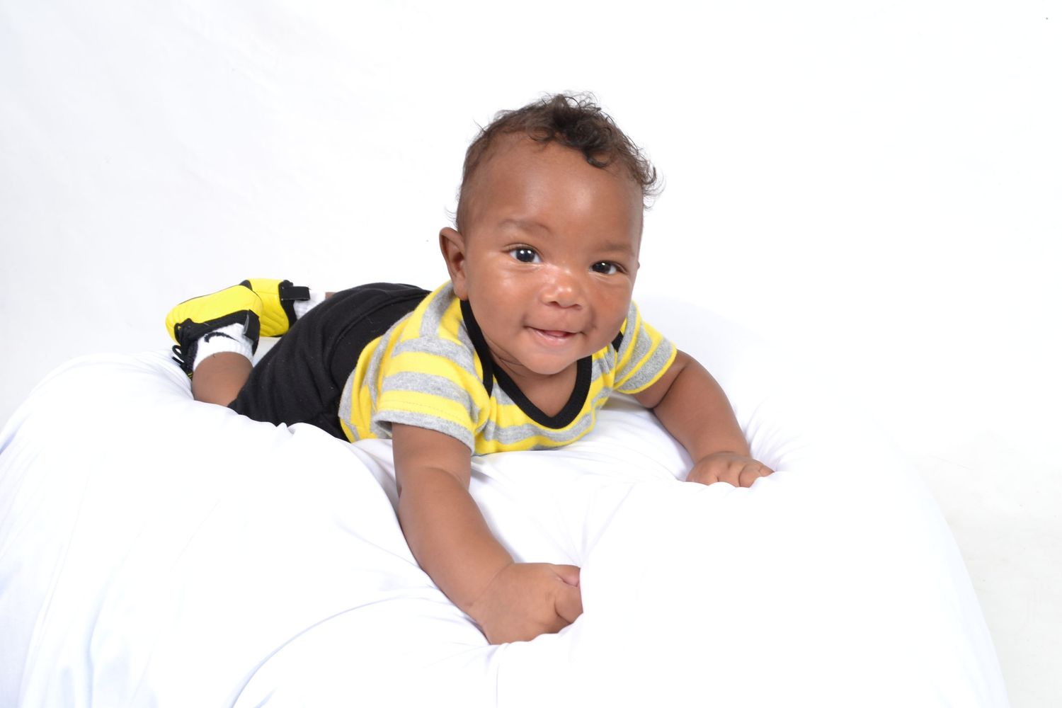 Adorable baby boy in striped yellow shirt lying on white surface smiling at camera.