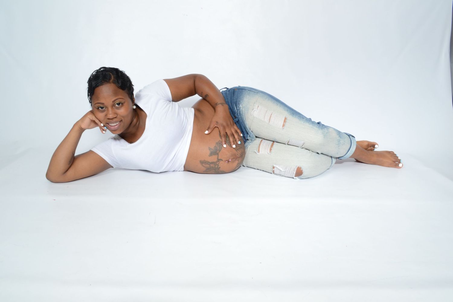 Pregnant woman with belly tattoos lies on white floor wearing jeans and white top, smiling.