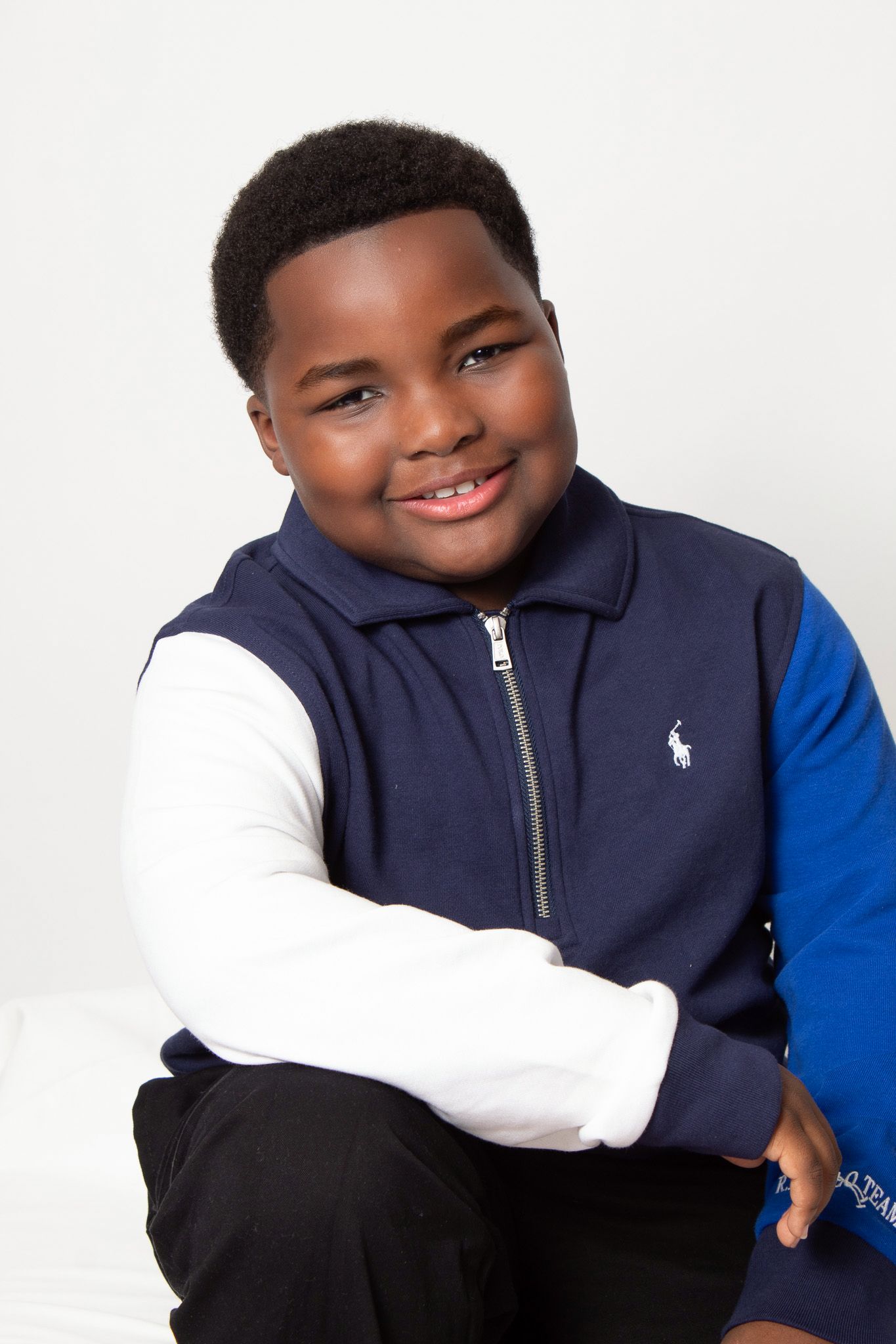 Smiling young boy wearing a navy blue Ralph Lauren zip-up jacket with white sleeves, sitting with arms crossed.
