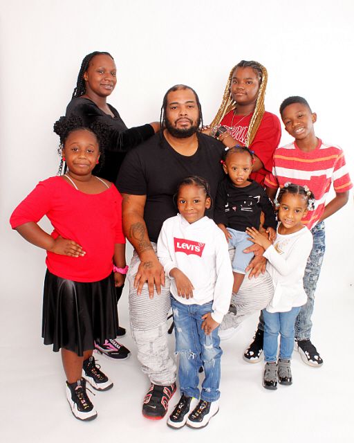 A family portrait of eight people, including adults and children, posing together in a white studio setting.